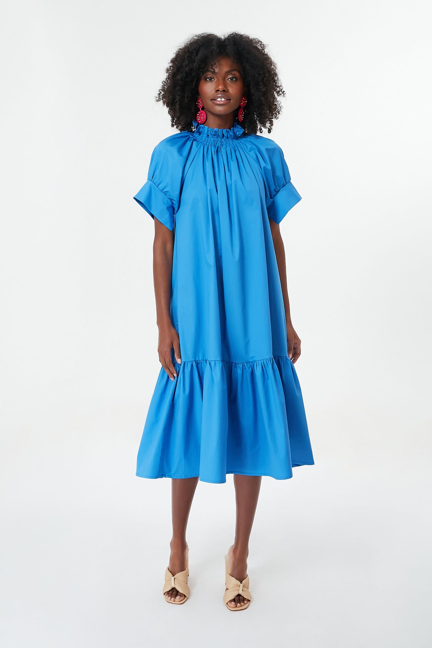 A woman wears a Monica Nera loose blue midi dress made from sustainable fabrics, featuring a ruffled high neckline, short sleeves, and hem, styled with beige heeled sandals and red statement earrings.