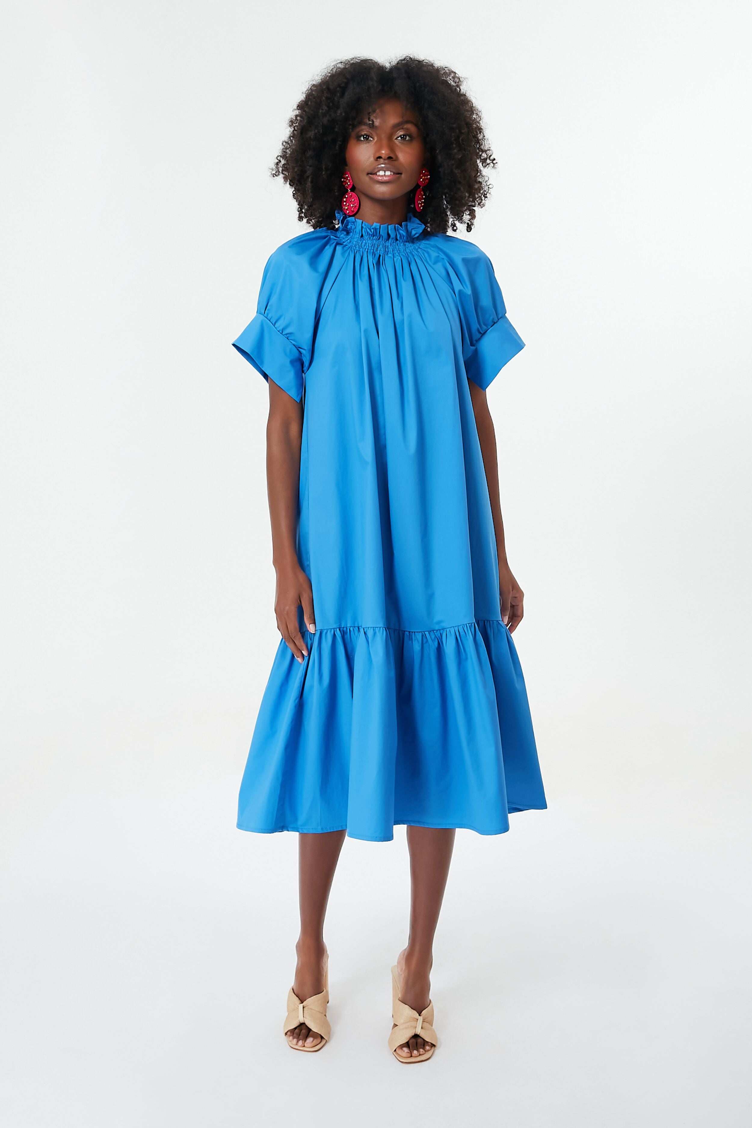 A woman wears a Monica Nera loose blue midi dress made from sustainable fabrics, featuring a ruffled high neckline, short sleeves, and hem, styled with beige heeled sandals and red statement earrings.