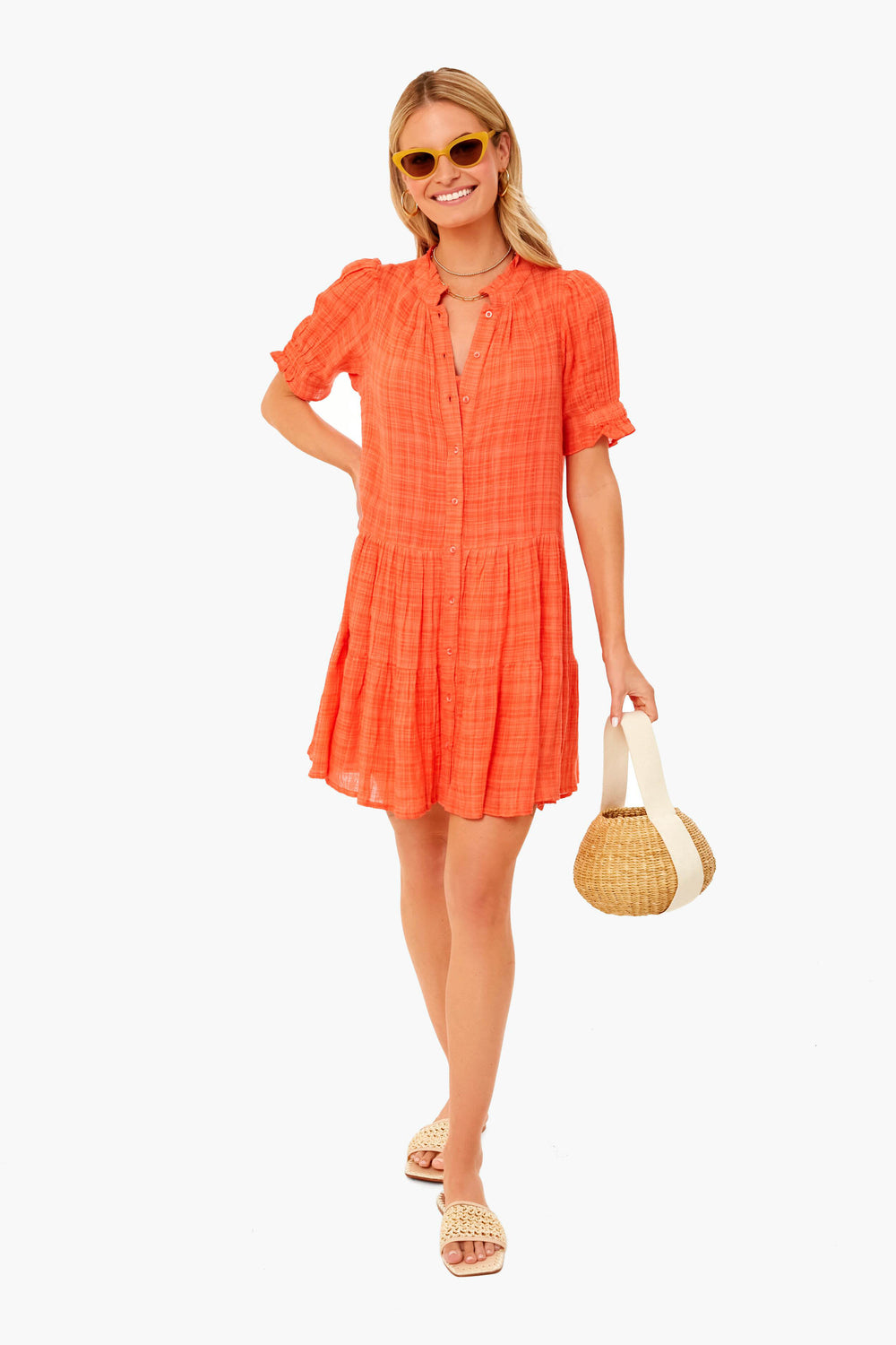 A woman wears an Apiece Apart short-sleeved orange plaid mini dress with a ruffle neck, yellow sunglasses, sandals, and holds a round straw bag.