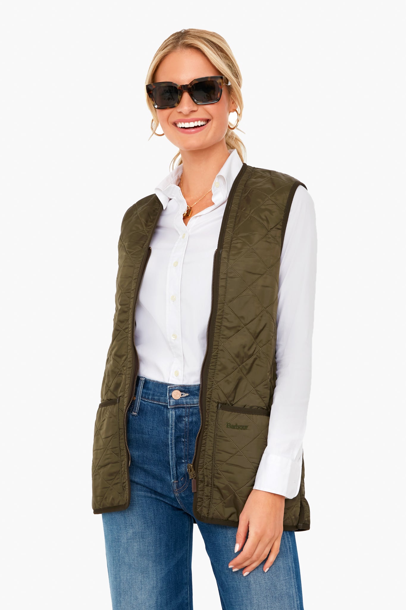 Smiling woman in sunglasses, a white button-down shirt, blue jeans, and an olive green Barbour quilted vest.