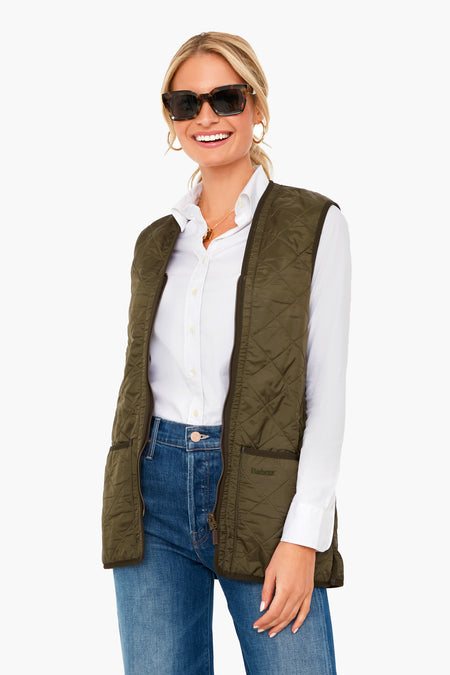 Smiling woman in sunglasses, a white button-down shirt, blue jeans, and an olive green Barbour quilted vest.