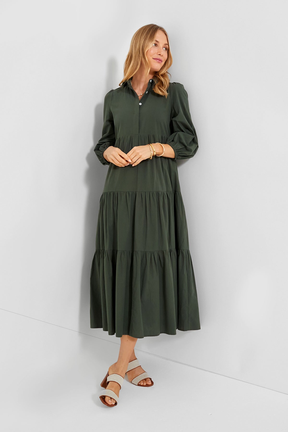 A woman wears a Pomander Place dark green cotton dress with long sleeves, button details, and a tiered midi silhouette, paired with light sandals and gold bracelets for an elegant look.