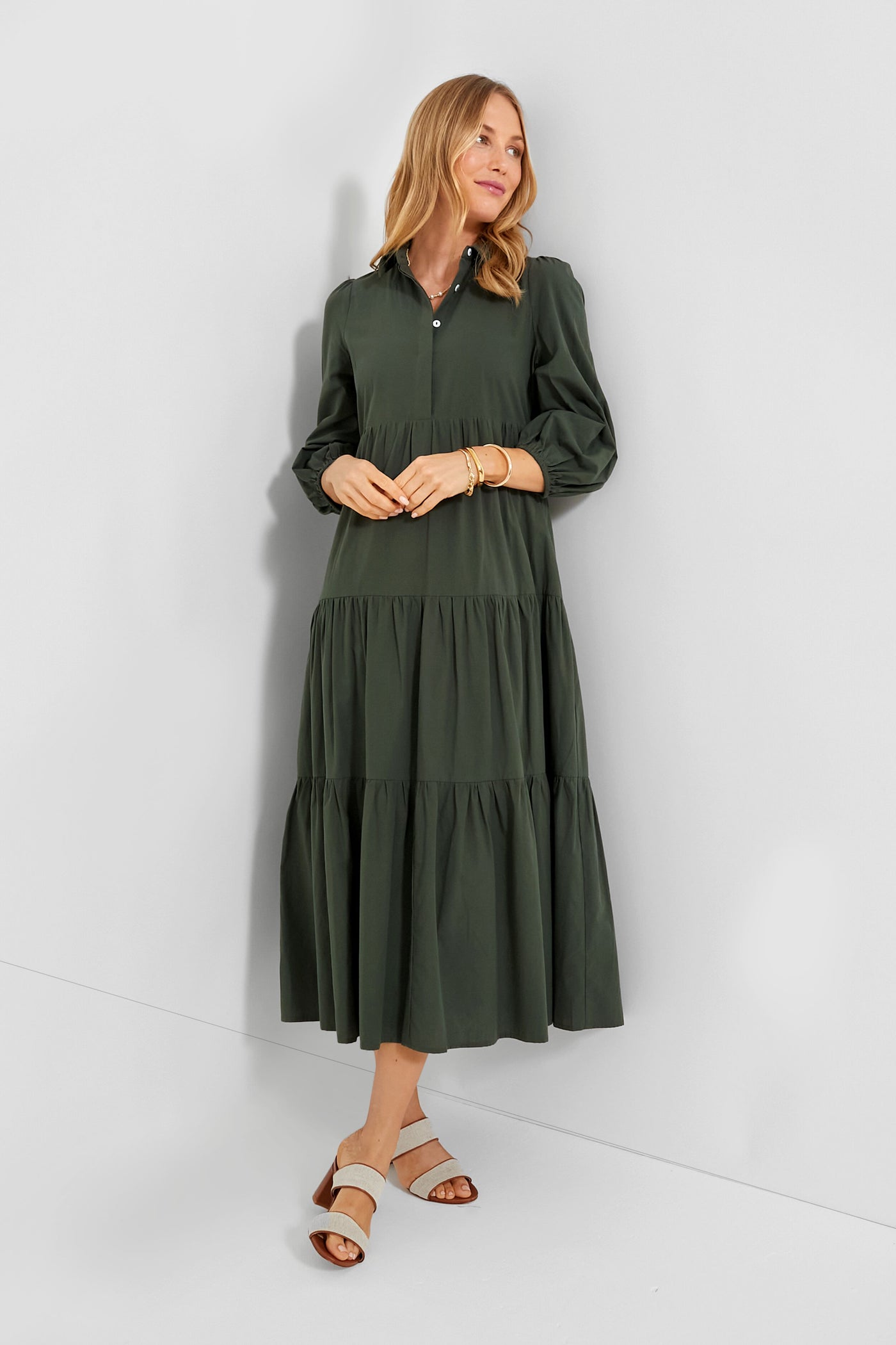 A woman wears a Pomander Place dark green cotton dress with long sleeves, button details, and a tiered midi silhouette, paired with light sandals and gold bracelets for an elegant look.
