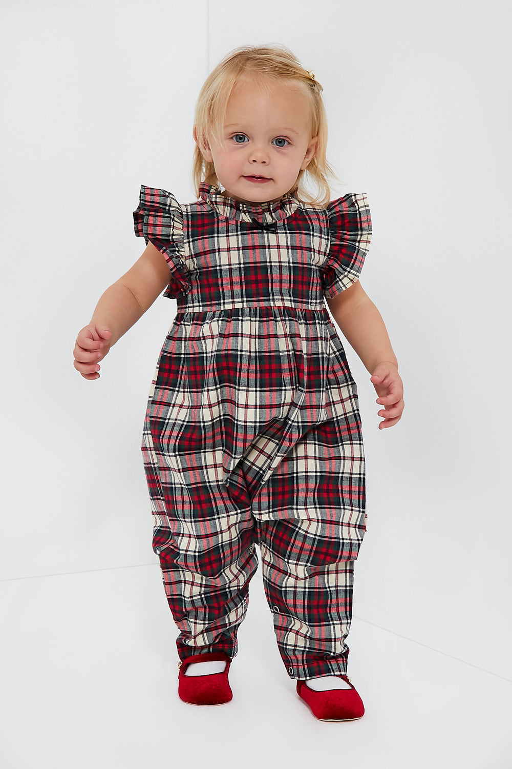 A toddler wears a Pink Chicken Jennifer Jumper made of brushed cotton with red, white, and black plaid, ruffled sleeves and neckline, paired with red shoes.