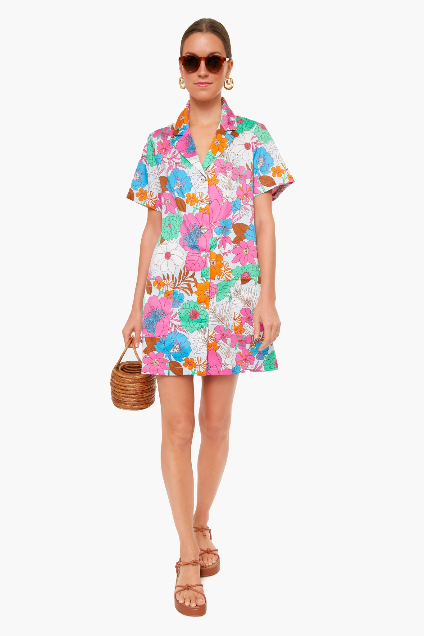 A woman wears a Tuckernuck Wild Rhapsody Townes Dress with colorful floral prints, brown sandals, large sunglasses, gold hoop earrings, and carries a small round wicker handbag—perfect for warm-weather outings.