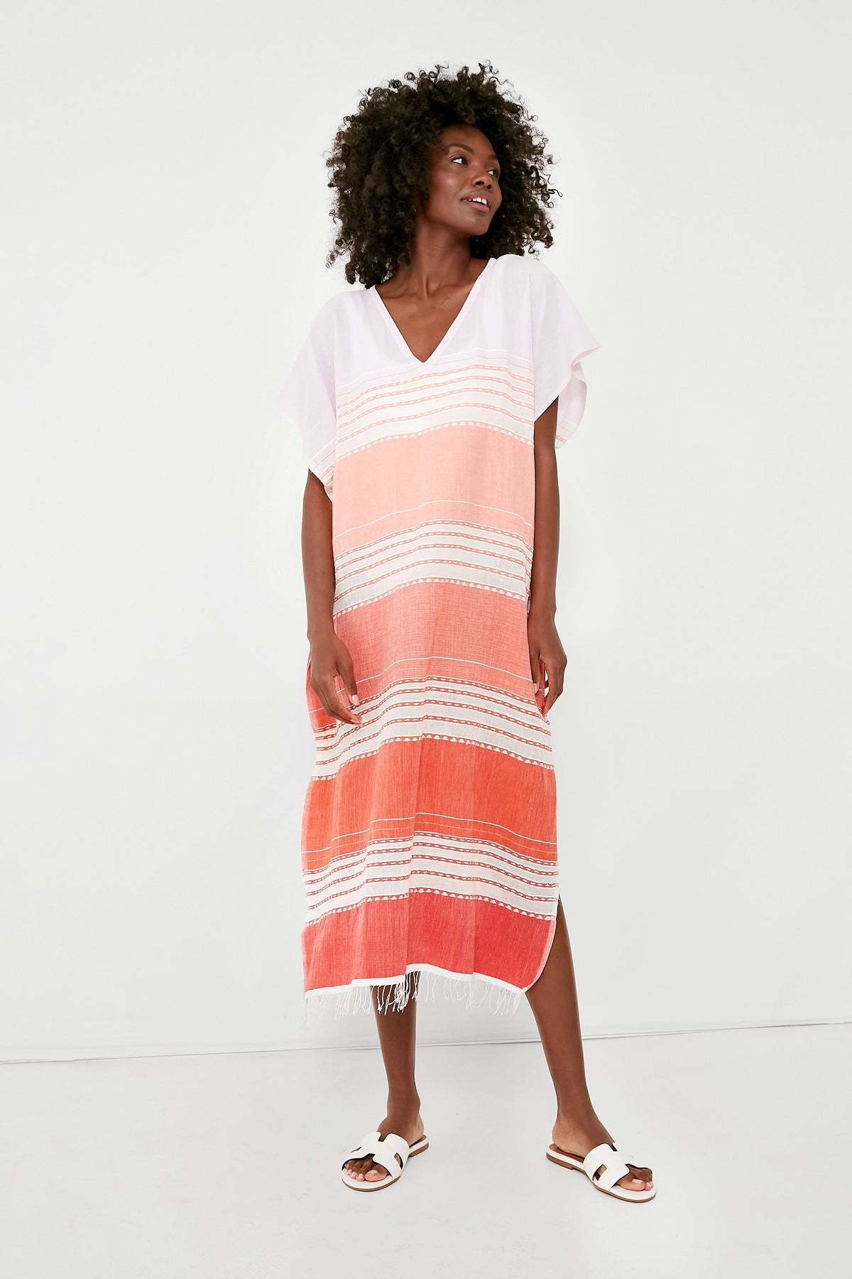 A woman wears a loose, short-sleeve, handwoven striped dress in pink and orange by lemlem, paired with white sandals.
