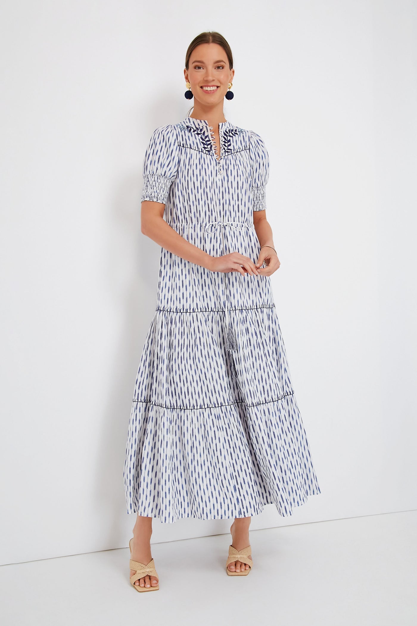 A woman wears the Carolina K Ikat Tara Dress, a white and blue organic cotton maxi with short sleeves and a tiered skirt, complemented by beige sandals and navy statement earrings.