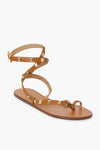 The Schutz tan flat sandal features a toe loop, ankle strap with buckle closure, gold square studs, and a simple leather sole—perfect for everyday wear.