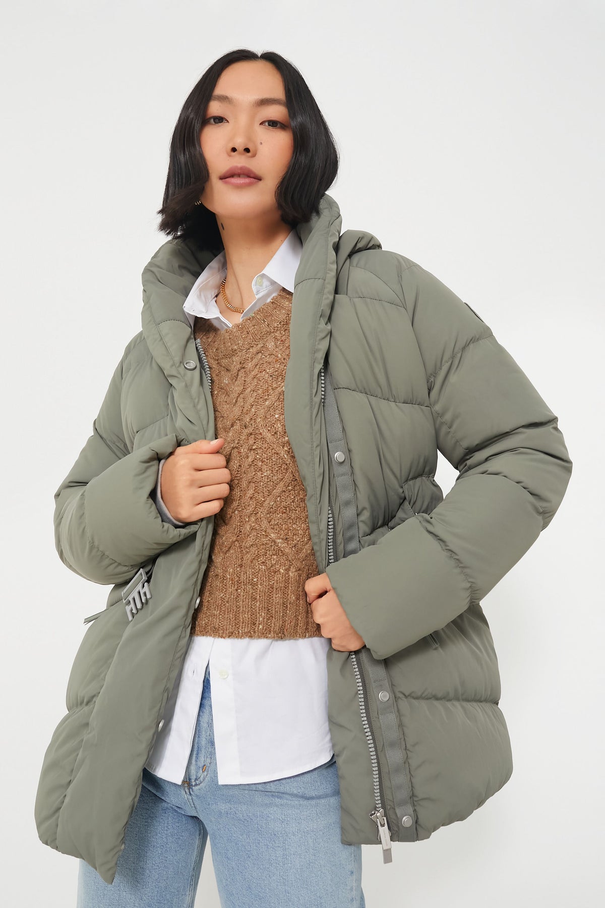 A person wears a Canada Goose Sagebrush Marlow Coat over a brown sweater, white collared shirt, and light blue jeans.