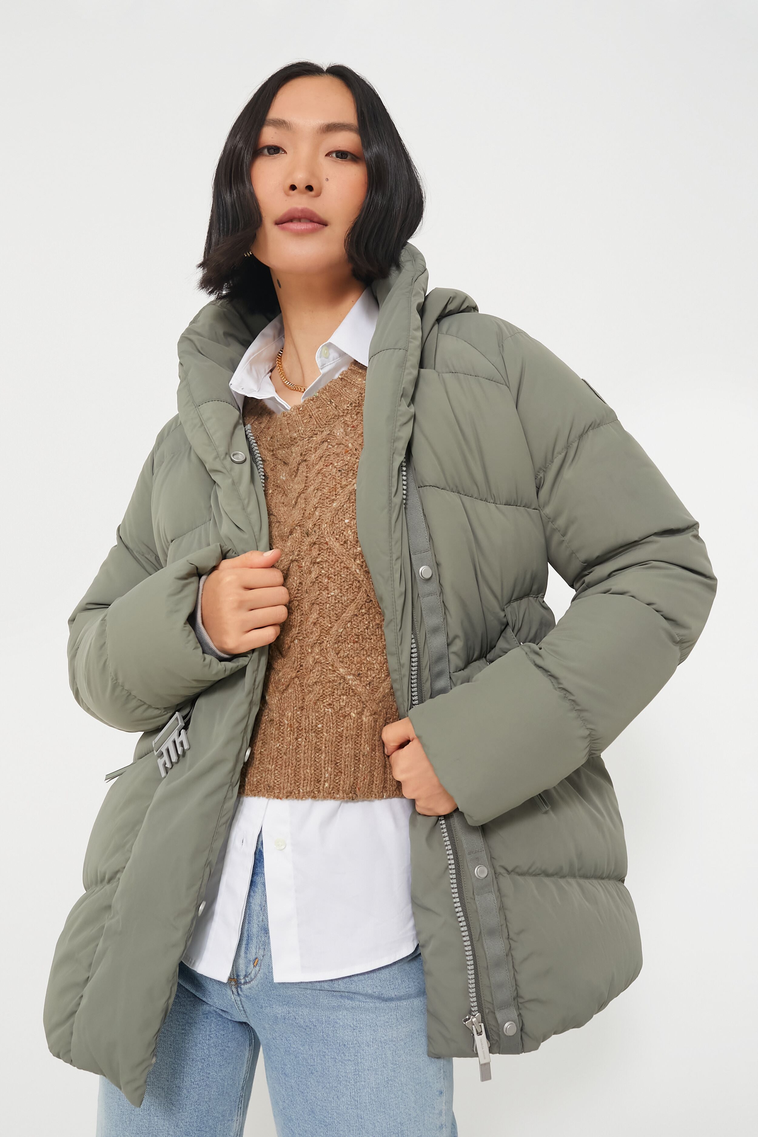 A person wears a Canada Goose Sagebrush Marlow Coat over a brown sweater, white collared shirt, and light blue jeans.