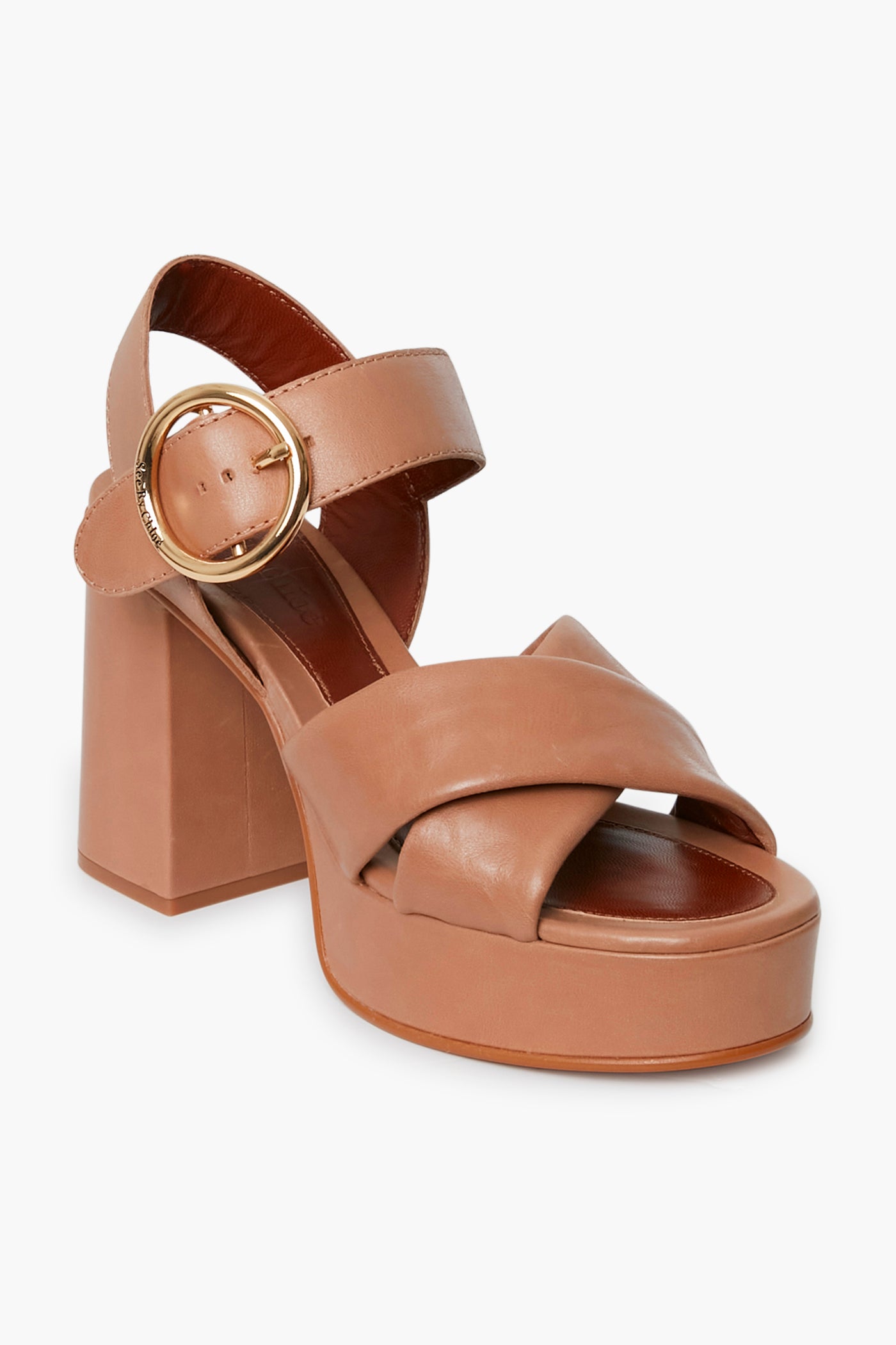 A tan platform sandal by See by Chloe featuring chunky high heel, wide crisscross front straps, and a large gold buckle ankle strap, inspired by the brand’s beige Lyna heels.