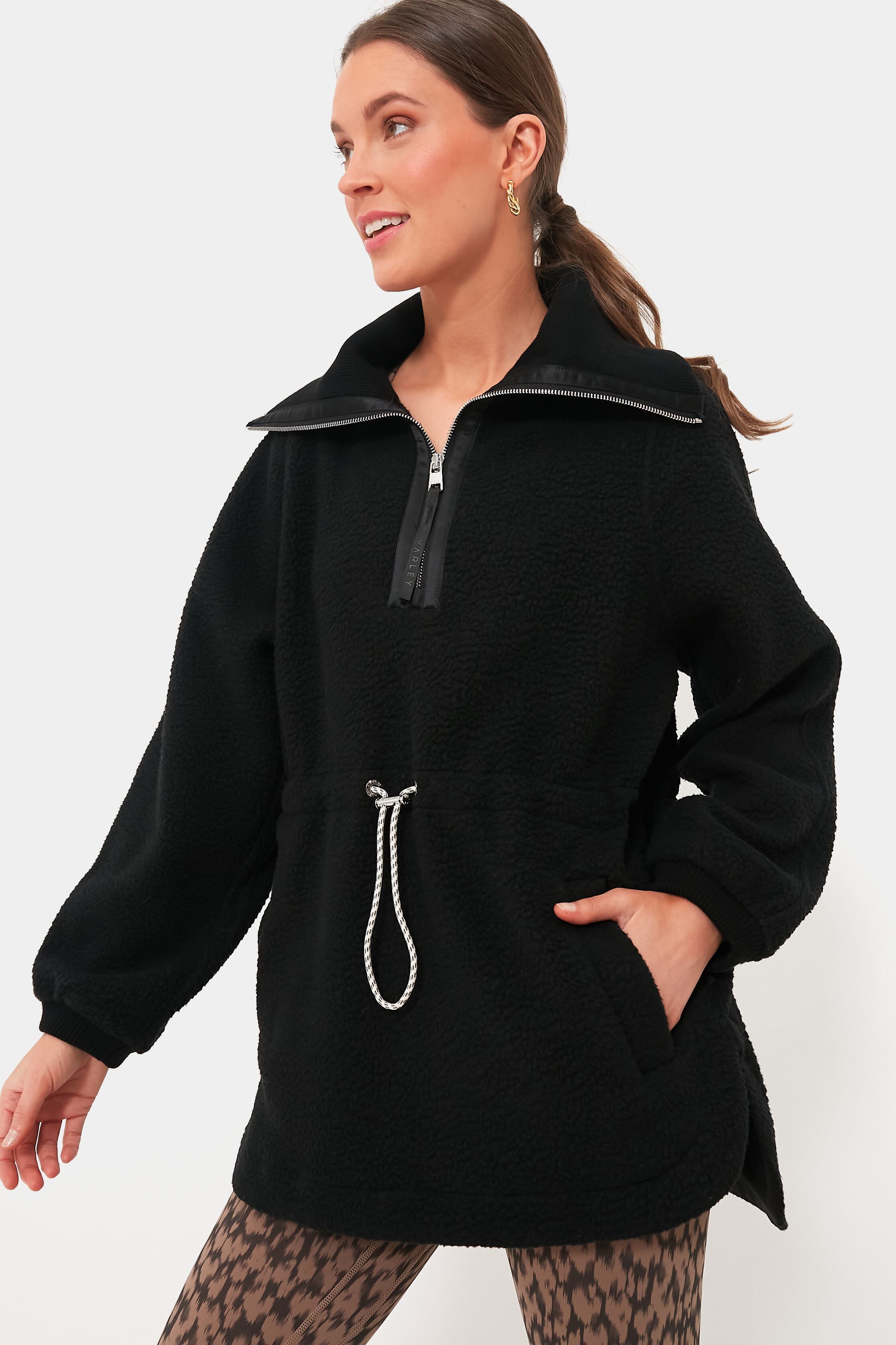 A woman wears a Varley black plush Borg fleece half-zip pullover with a drawstring waist, Alpine loungewear patterned leggings, and gold earrings.