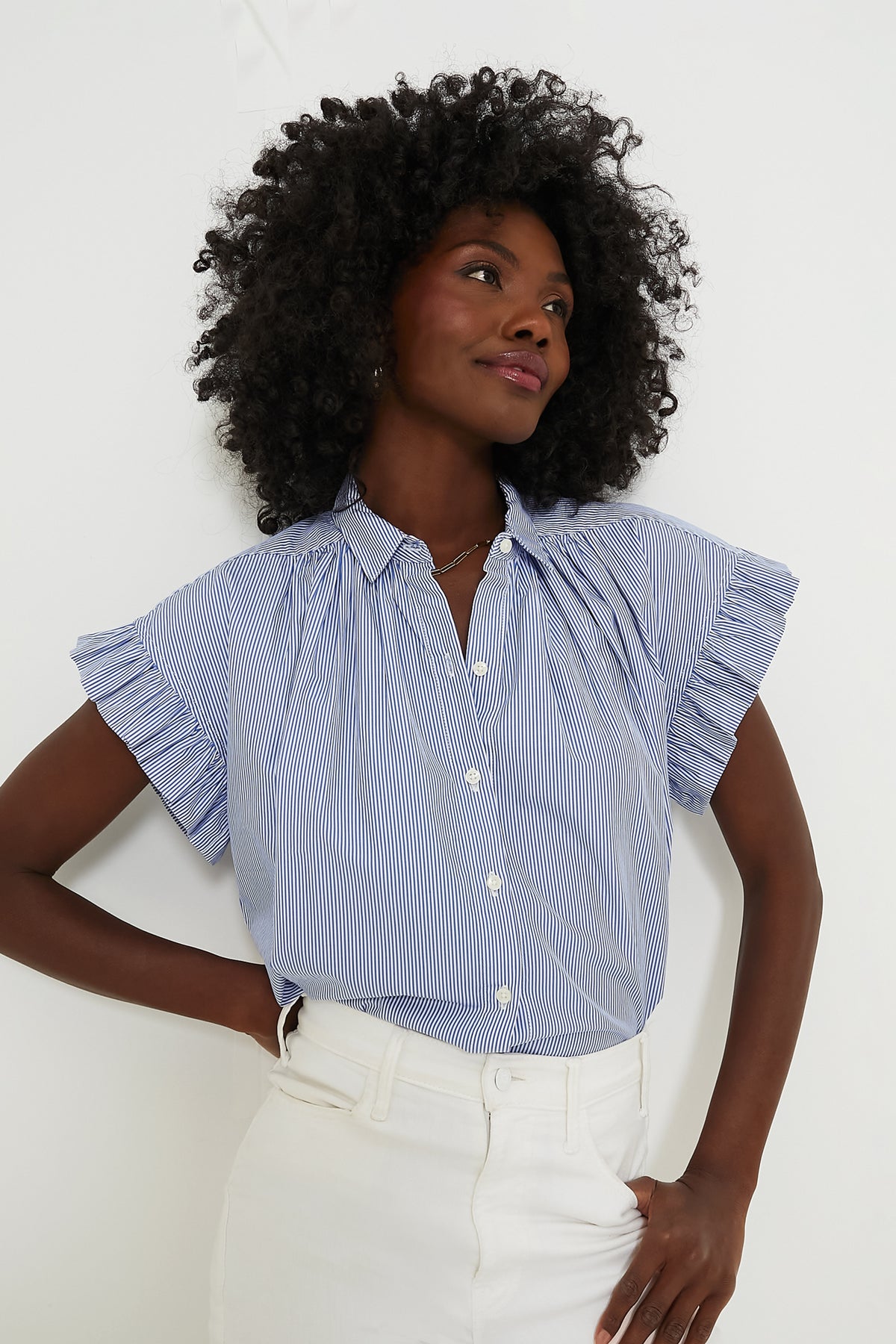 A woman wears a TROVATA Birds of Paradis blue and white striped short-sleeve blouse with ruffled sleeves, paired with white high-waisted pants.