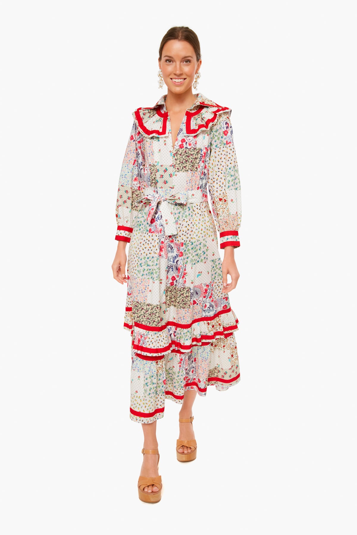A woman wears a Hunter Bell Jasper Dress in maxi length with floral patchwork prints and red accents, styled with long sleeves, a belt, tan platform sandals, and statement earrings.