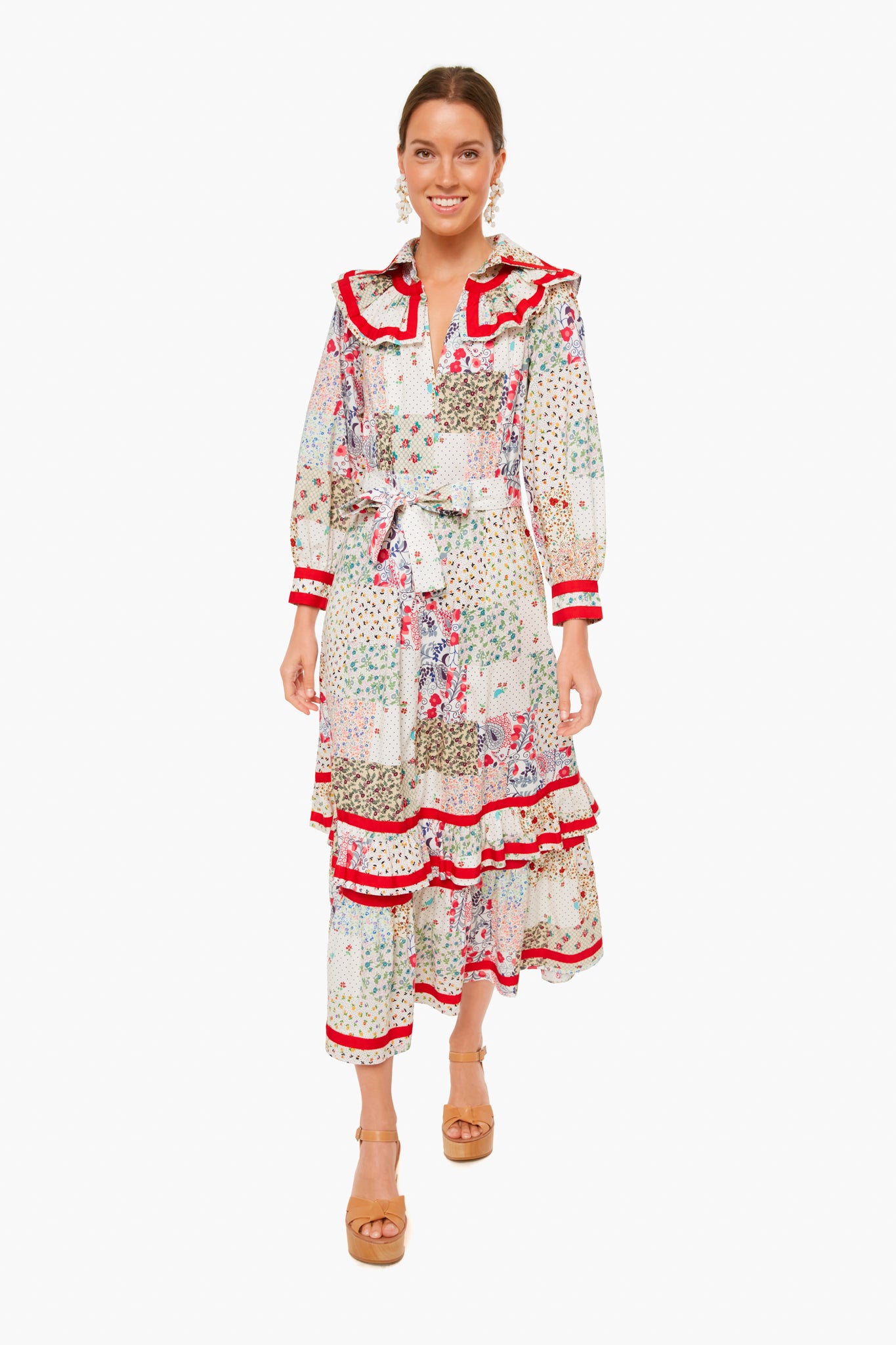 A woman wears a Hunter Bell Jasper Dress in maxi length with floral patchwork prints and red accents, styled with long sleeves, a belt, tan platform sandals, and statement earrings.