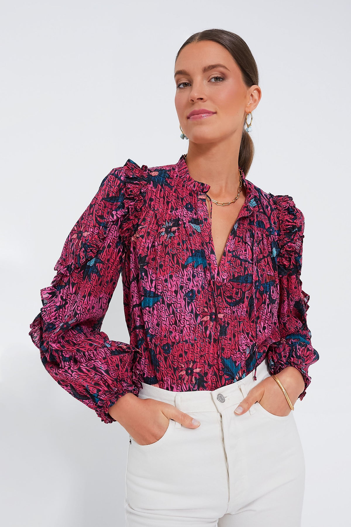 A woman wears the Ulla Johnson Manet Blouse, featuring long sleeves, floral print, and ruffle details, tucked into high-waisted white pants.