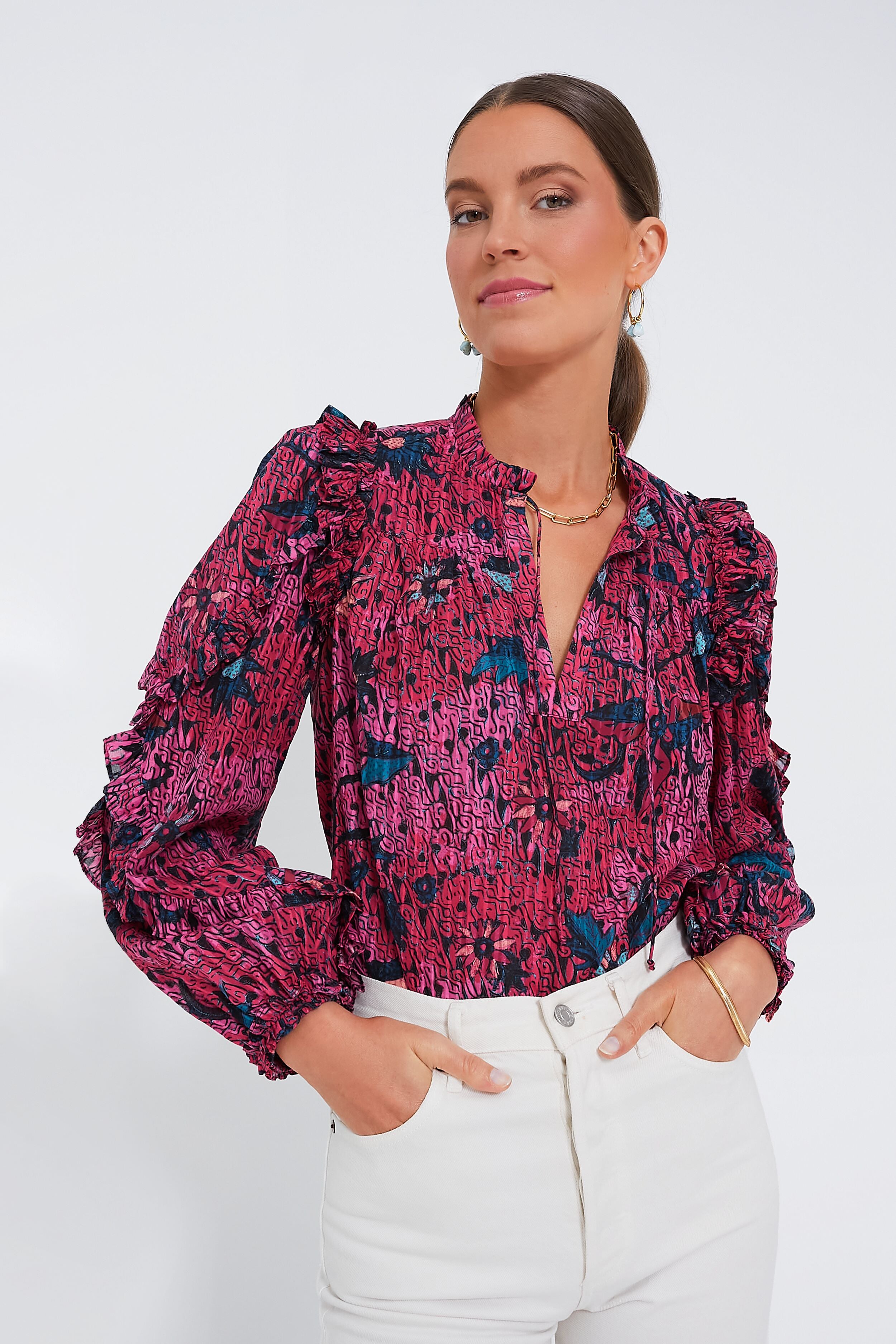 A woman wears the Ulla Johnson Manet Blouse, featuring long sleeves, floral print, and ruffle details, tucked into high-waisted white pants.