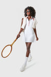 A woman in a Tnuck Sport white tennis dress and sunglasses holds a wooden racket, sweater draped over her shoulders, standing confidently.