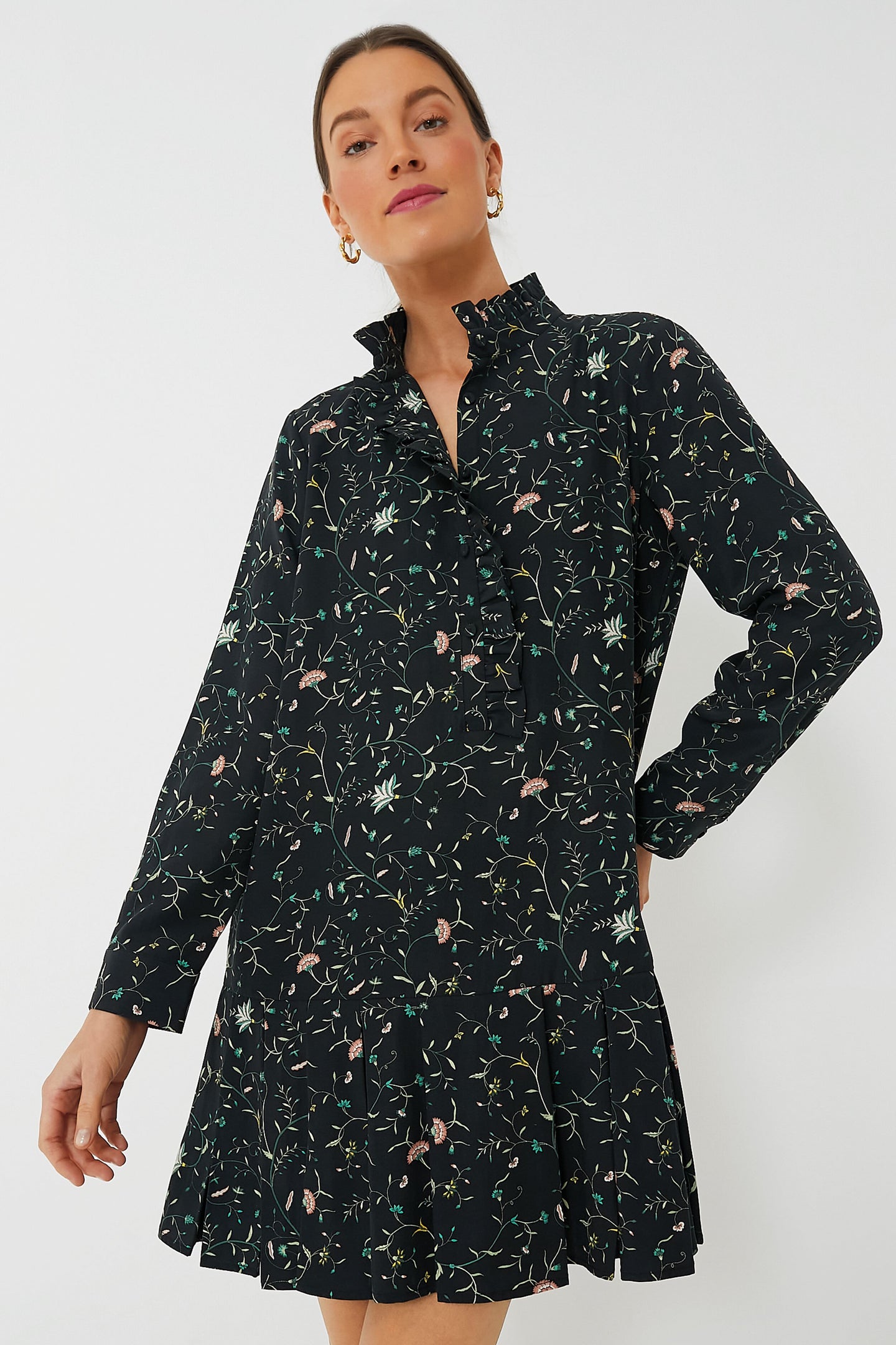 A woman stands in a simple setting wearing a Tuckernuck long-sleeve black floral shirt dress with a ruffle collar.