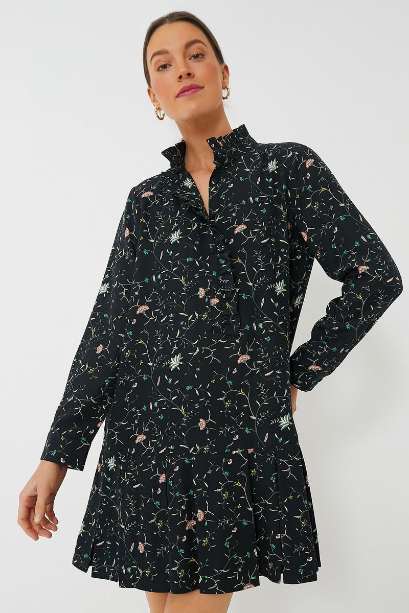 A woman stands in a simple setting wearing a Tuckernuck long-sleeve black floral shirt dress with a ruffle collar.