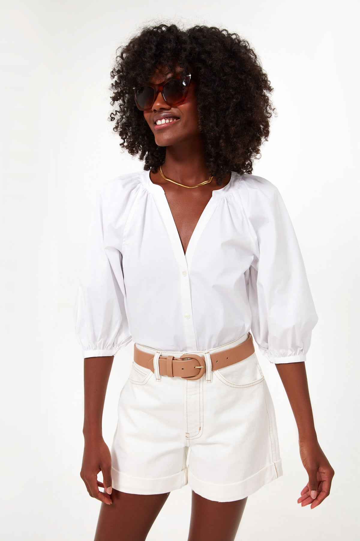 A woman wears STAUD sunglasses, a cotton poplin puff-sleeve blouse, and high-waisted white shorts with a beige belt.