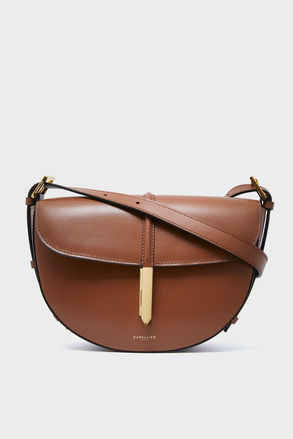 The Demellier Tan Tokyo Saddle Bag is a brown leather crossbody featuring a curved flap, gold-tone hardware, and an adjustable strap—an elegant example of refined handcrafted style.