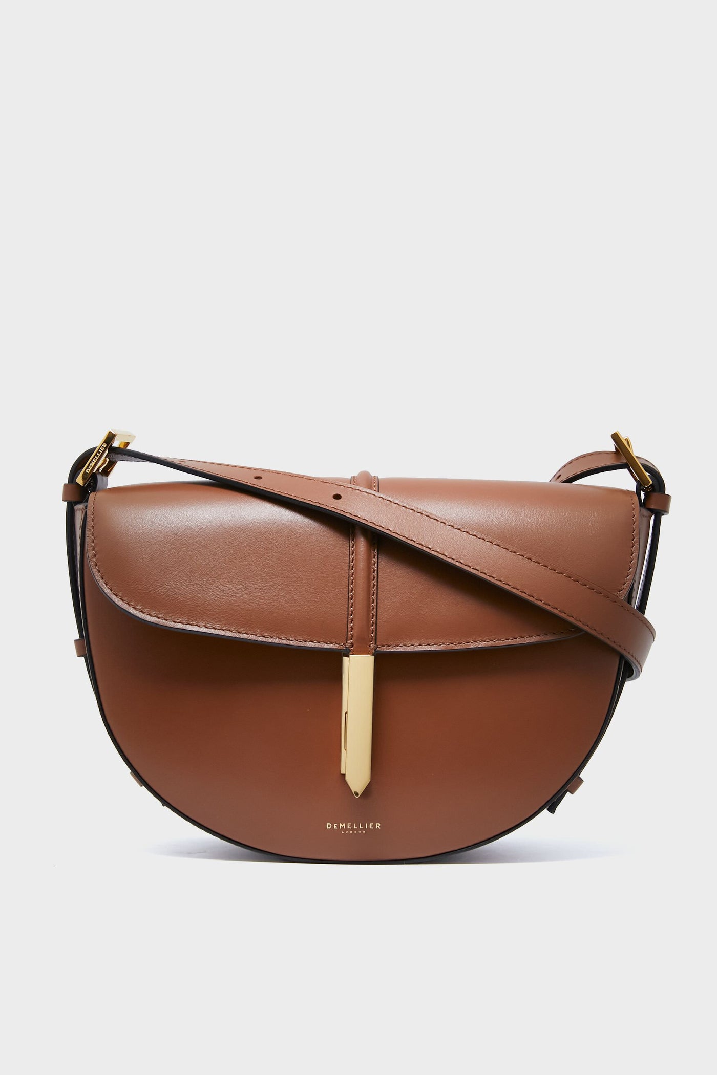 The Demellier Tan Tokyo Saddle Bag is a brown leather crossbody featuring a curved flap, gold-tone hardware, and an adjustable strap—an elegant example of refined handcrafted style.