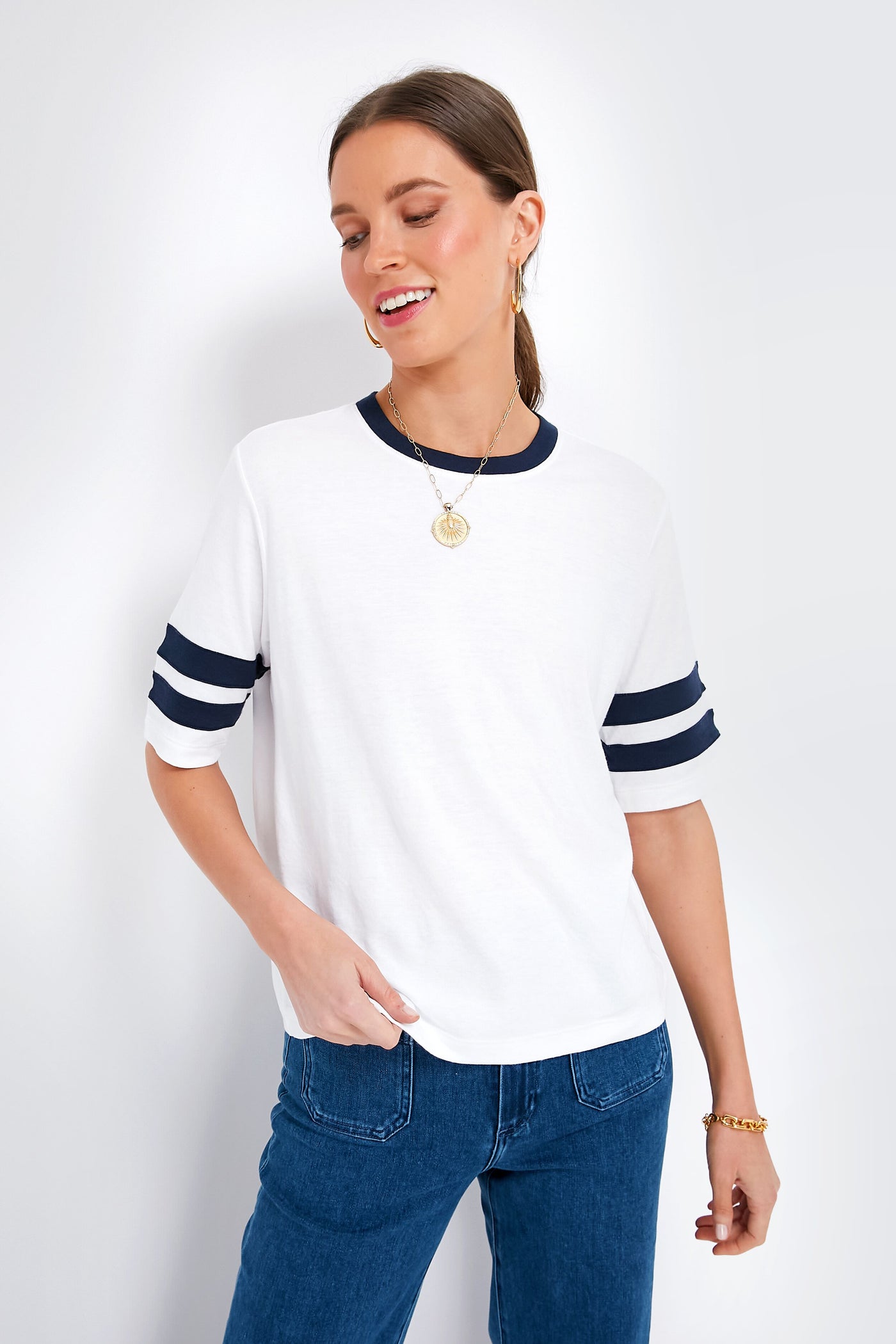A woman wears a Faherty varsity tee made from soft pima cotton, featuring a white body with navy collar and sleeve stripes, styled with blue jeans, a gold necklace, and bracelet.