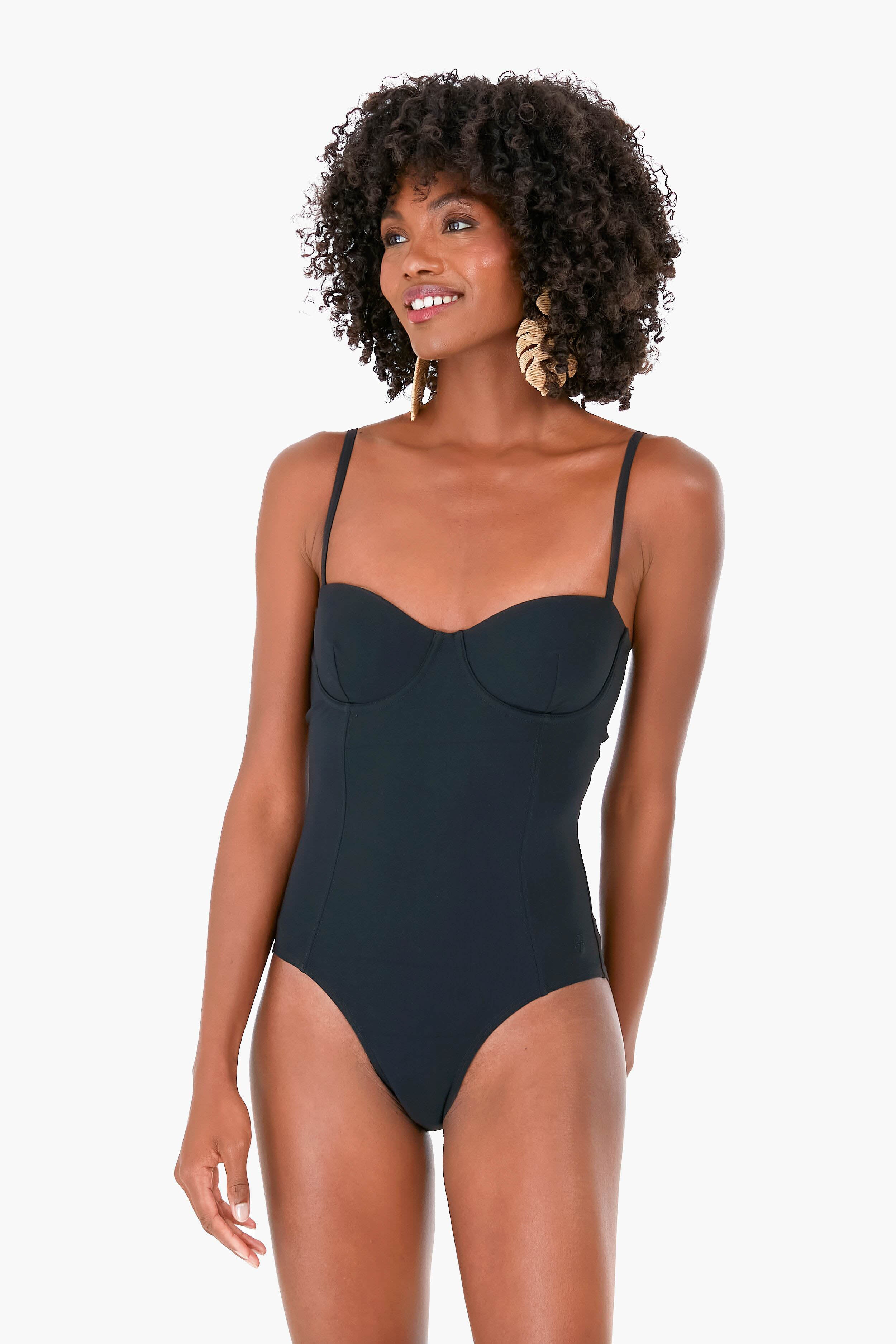 A woman stands confidently wearing a Tory Burch black underwire one-piece swimsuit with thin straps.