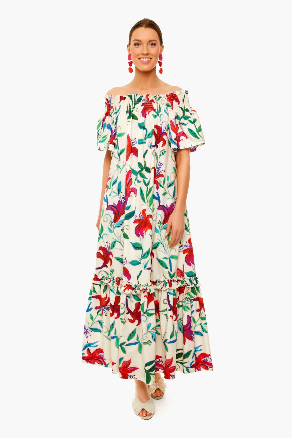 A woman wears a La DoubleJ Capalbio Dress in vibrant floral cotton poplin, styled with red statement earrings and white open-toe shoes.