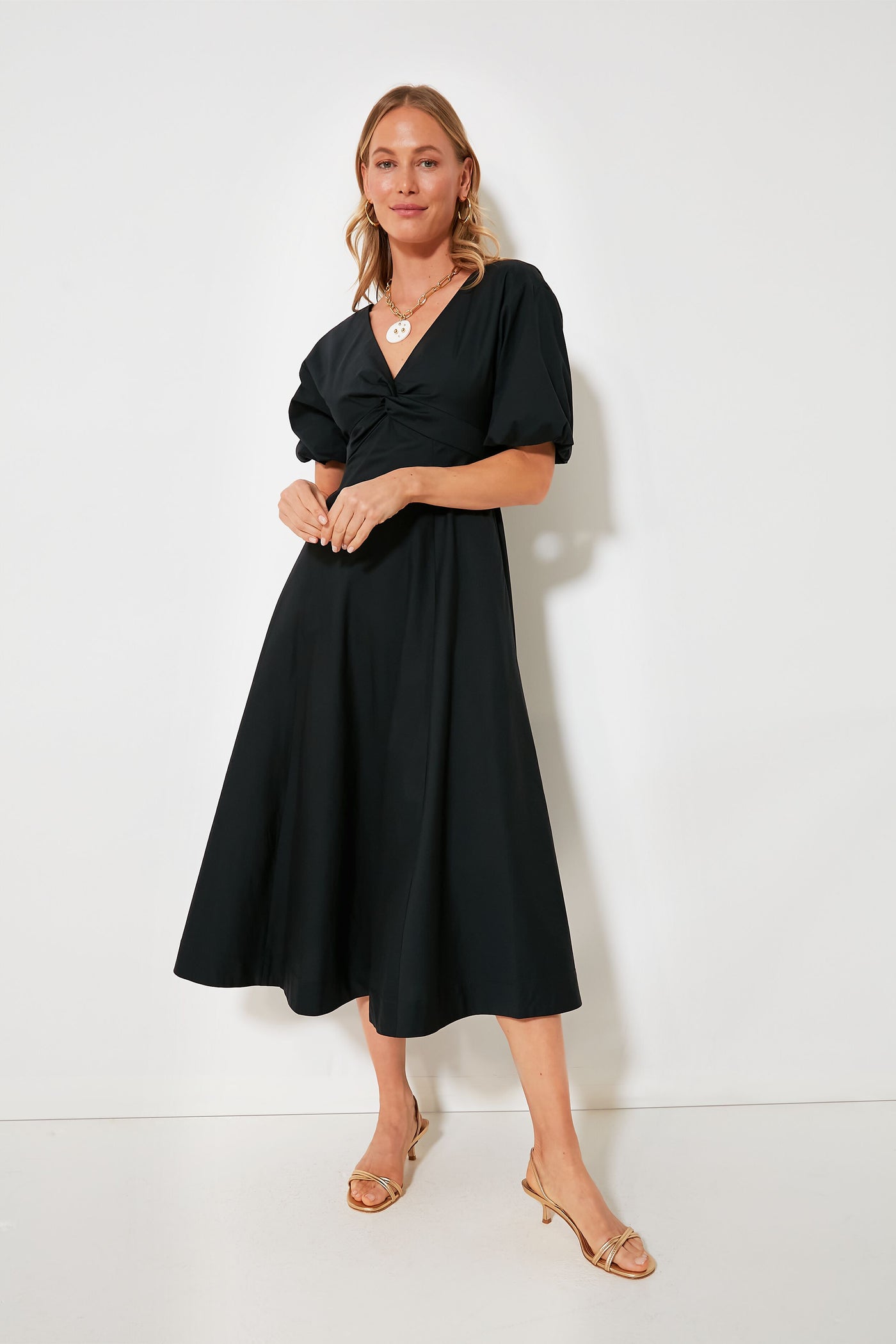 A woman wears the STAUD Black Finley Dress, a cotton poplin midi with elegant puff sleeves, paired with gold open-toe heels.