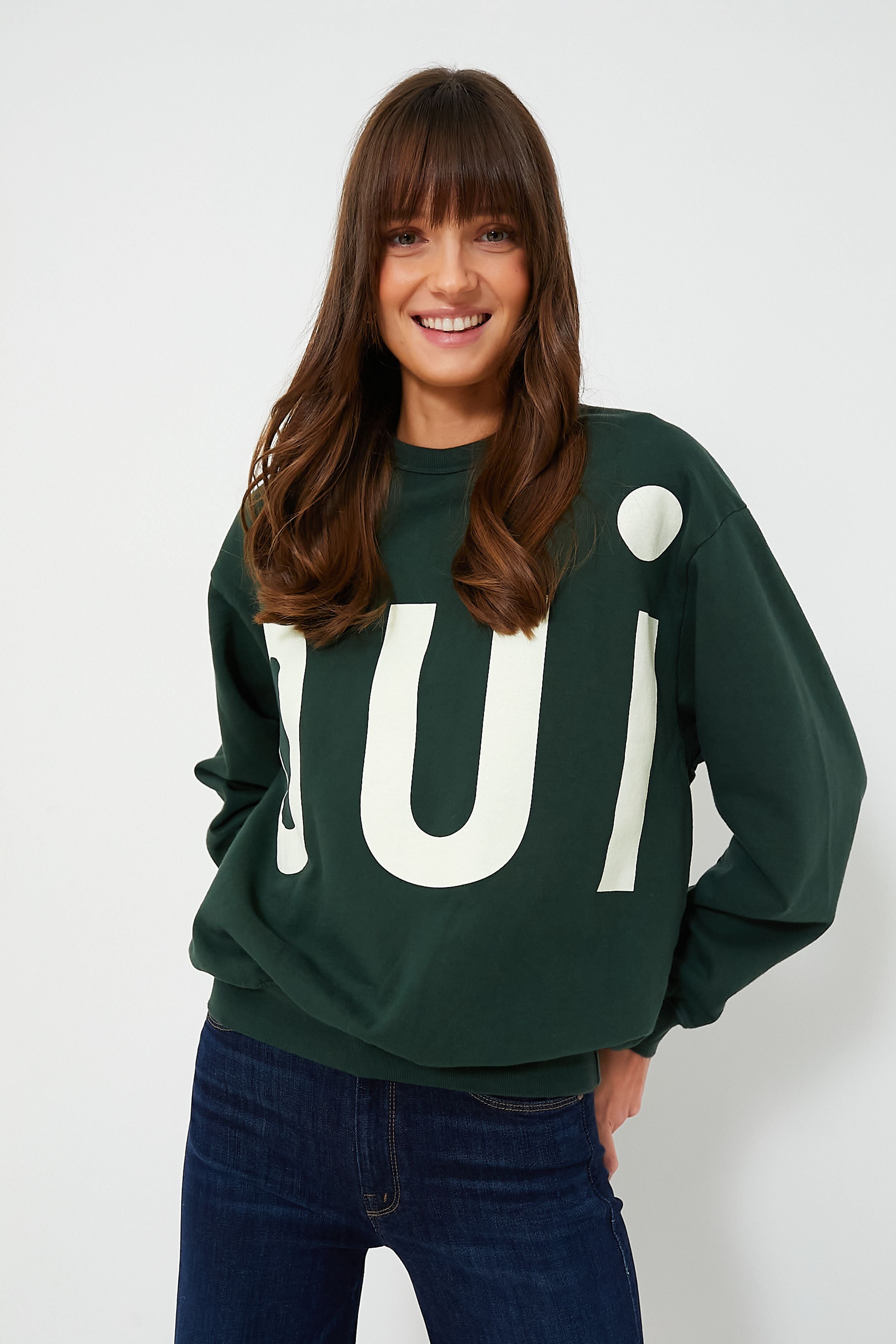 A woman with long brown hair wears a dark green Clare V Oui print sweater and dark blue jeans, with "Oui" in large white letters.