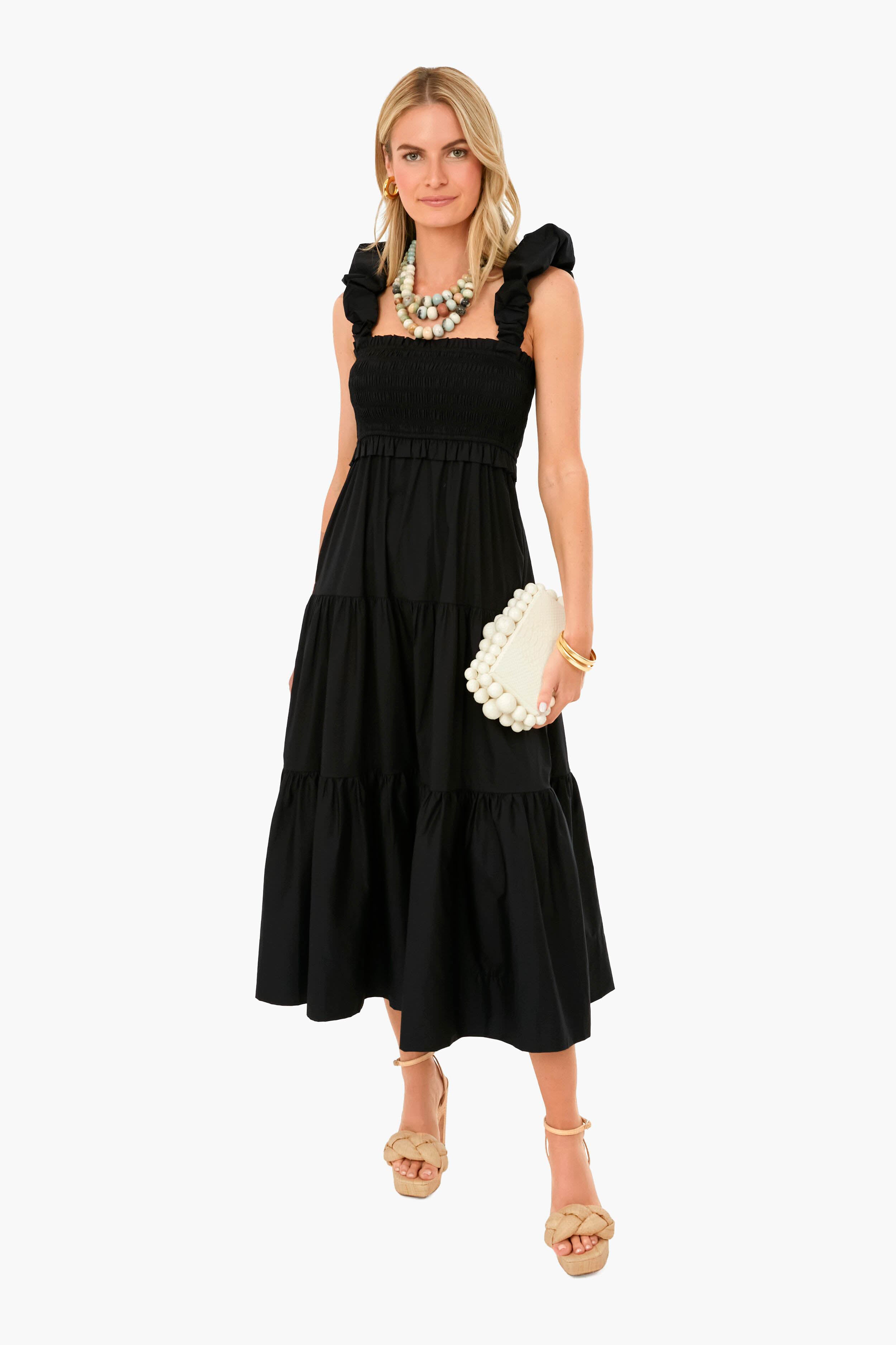 Woman in a Sea New York black sleeveless midi dress with ruffled straps, beige platform sandals, statement necklace, and carrying a cream clutch.