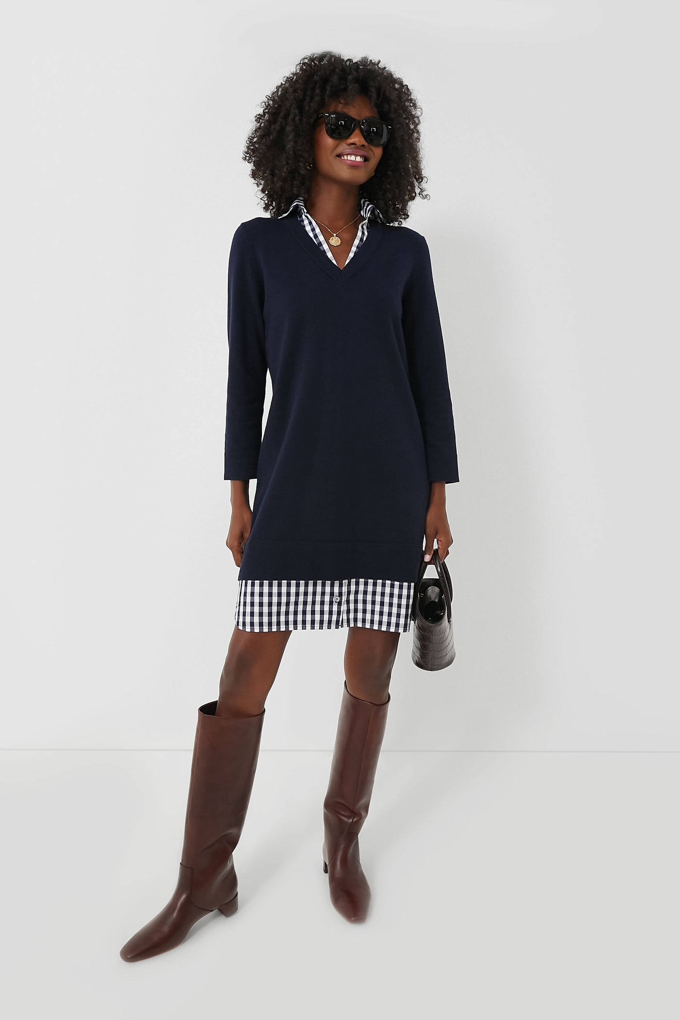 A woman in Southern style wears a Draper James Navy Wool Cotton Sweaterdress over a checkered shirt, paired with brown knee-high boots, sunglasses, and a small handbag.