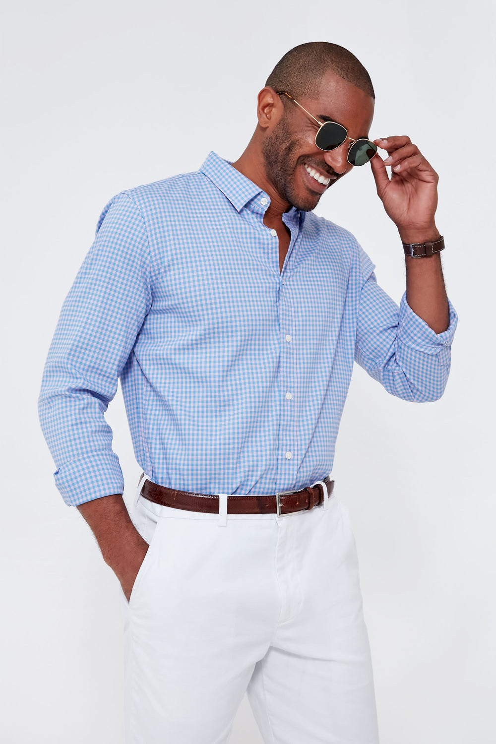 A man in Faherty sunglasses, a blue checked performance stretch shirt, white pants, and a brown belt stands with one hand in his pocket, smiling.