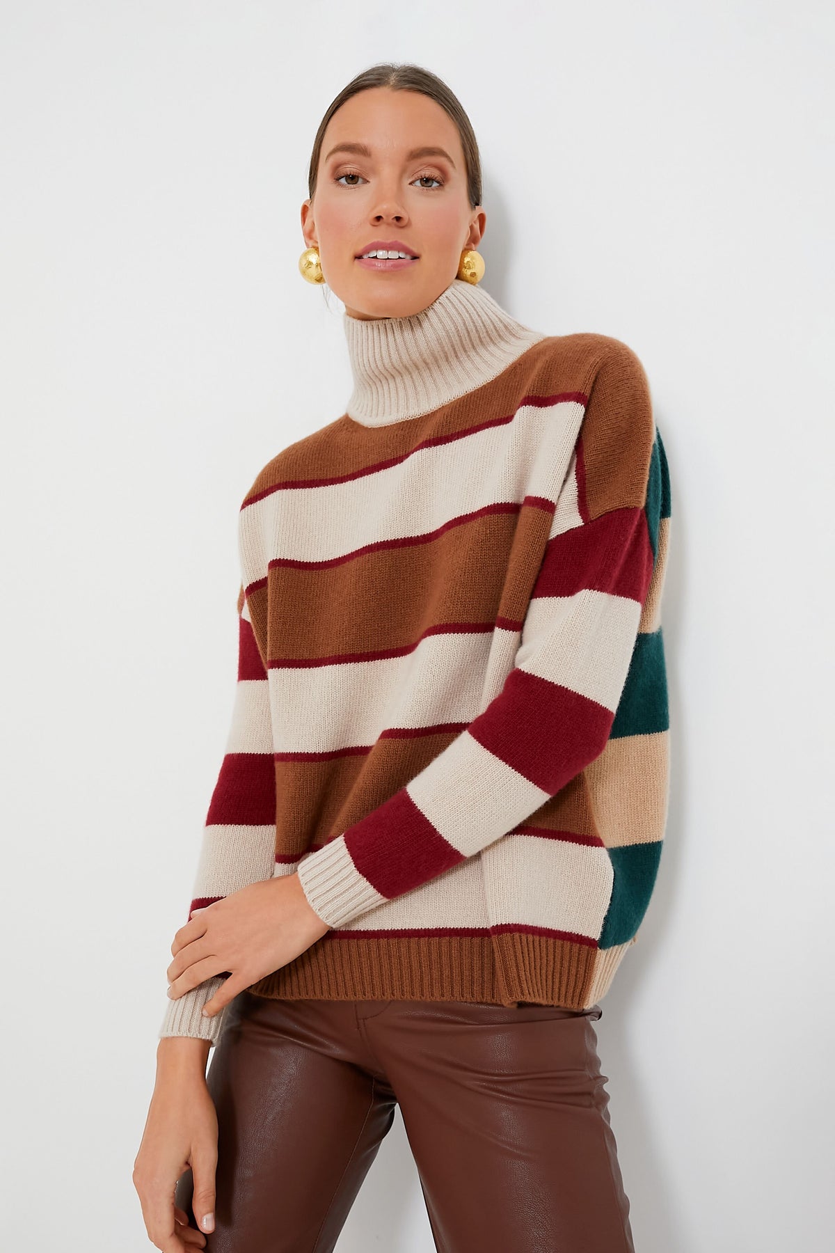 A person poses in a striped turtleneck virgin wool sweater by Weekend Max Mara in brown, cream, burgundy, and green tones, paired with brown leather pants—an ideal choice for fall-winter wardrobes.