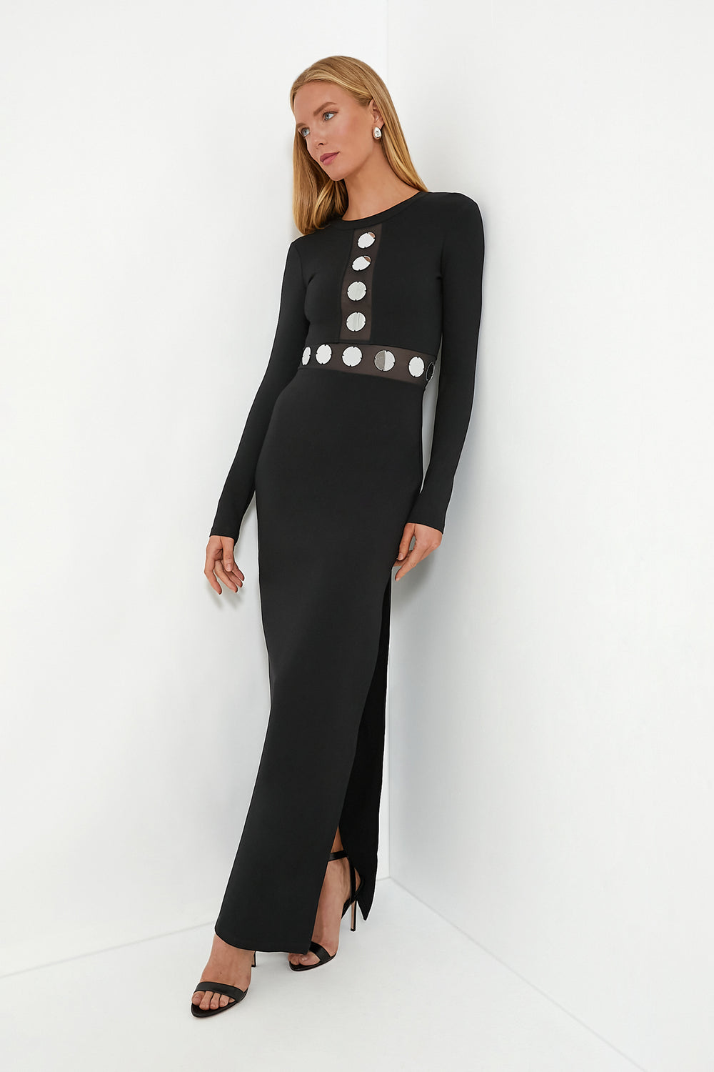 Woman in a STAUD black maxi dress with silver circular and mirrored details, featuring a side slit, paired with black high heels and drop earrings.