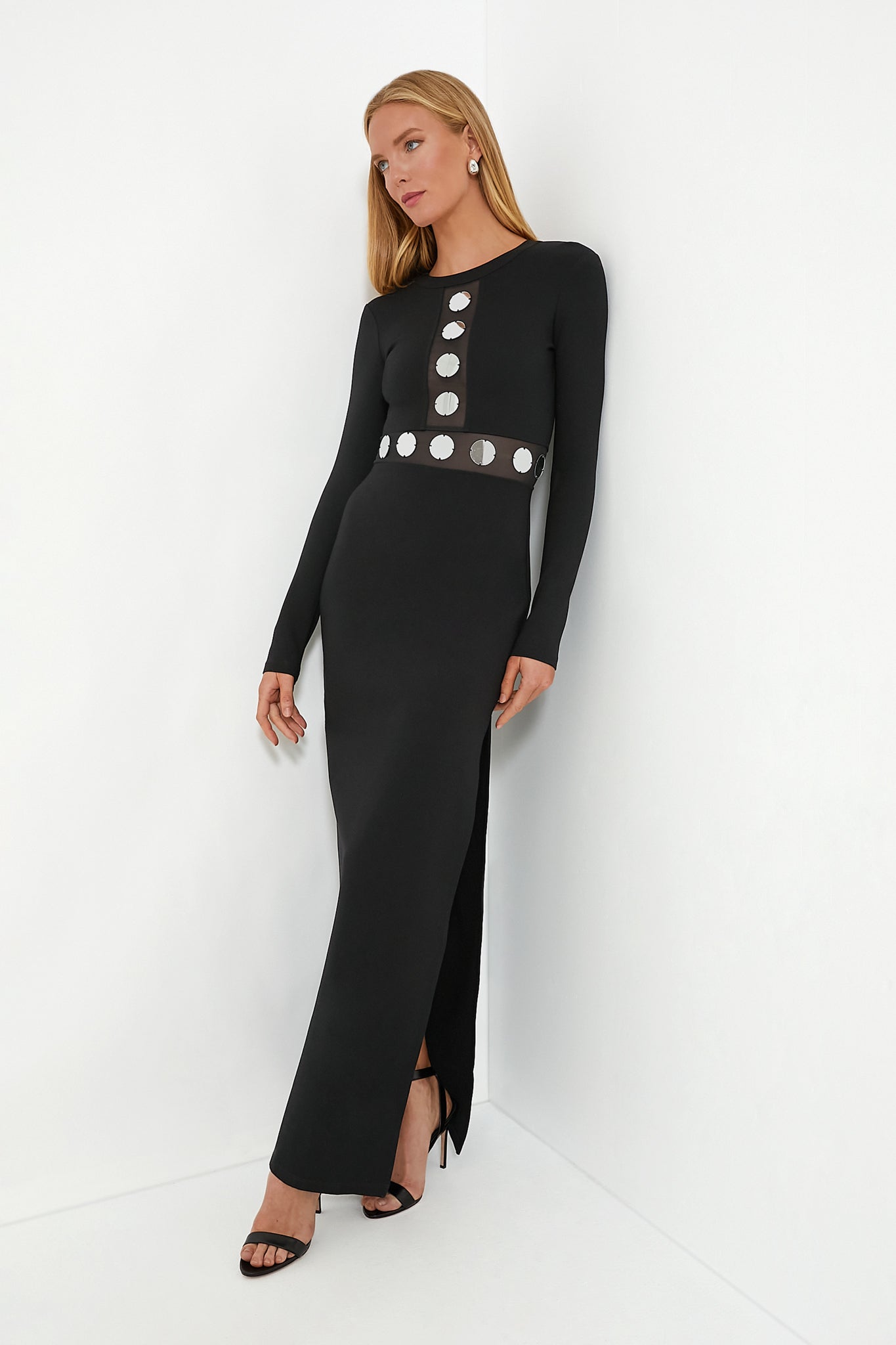 Woman in a STAUD black maxi dress with silver circular and mirrored details, featuring a side slit, paired with black high heels and drop earrings.