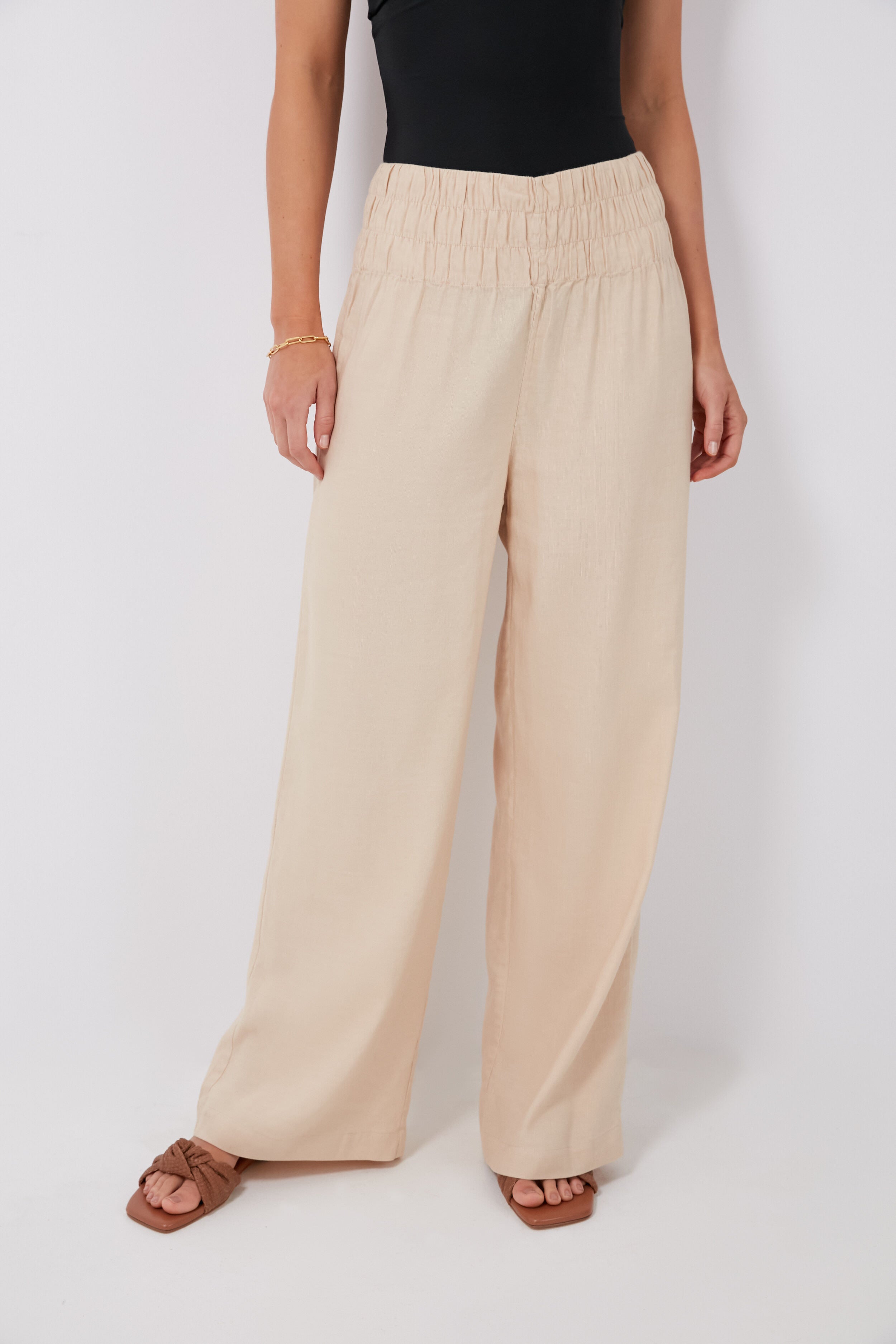 A person confidently stands in onia high-waisted, wide-leg Sand Air Smocked Pants (linen blend), paired with a black sleeveless top, brown sandals, and a gold bracelet.