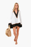 A woman smiles confidently in a Caroline Constas white linen coverup with black trim, paired with a woven bag with wooden handles and tan sandals, showcasing chic designer beachwear.