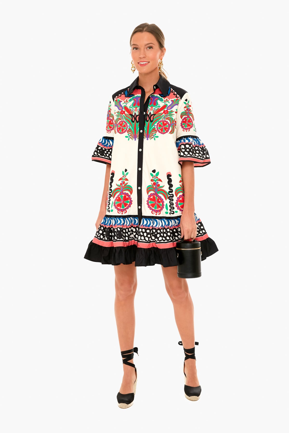 A woman wears a La DoubleJ cotton poplin dress with vibrant floral and geometric prints, black ruffled hem, paired with black espadrille wedges and a small black cylindrical handbag. Made in Italy.