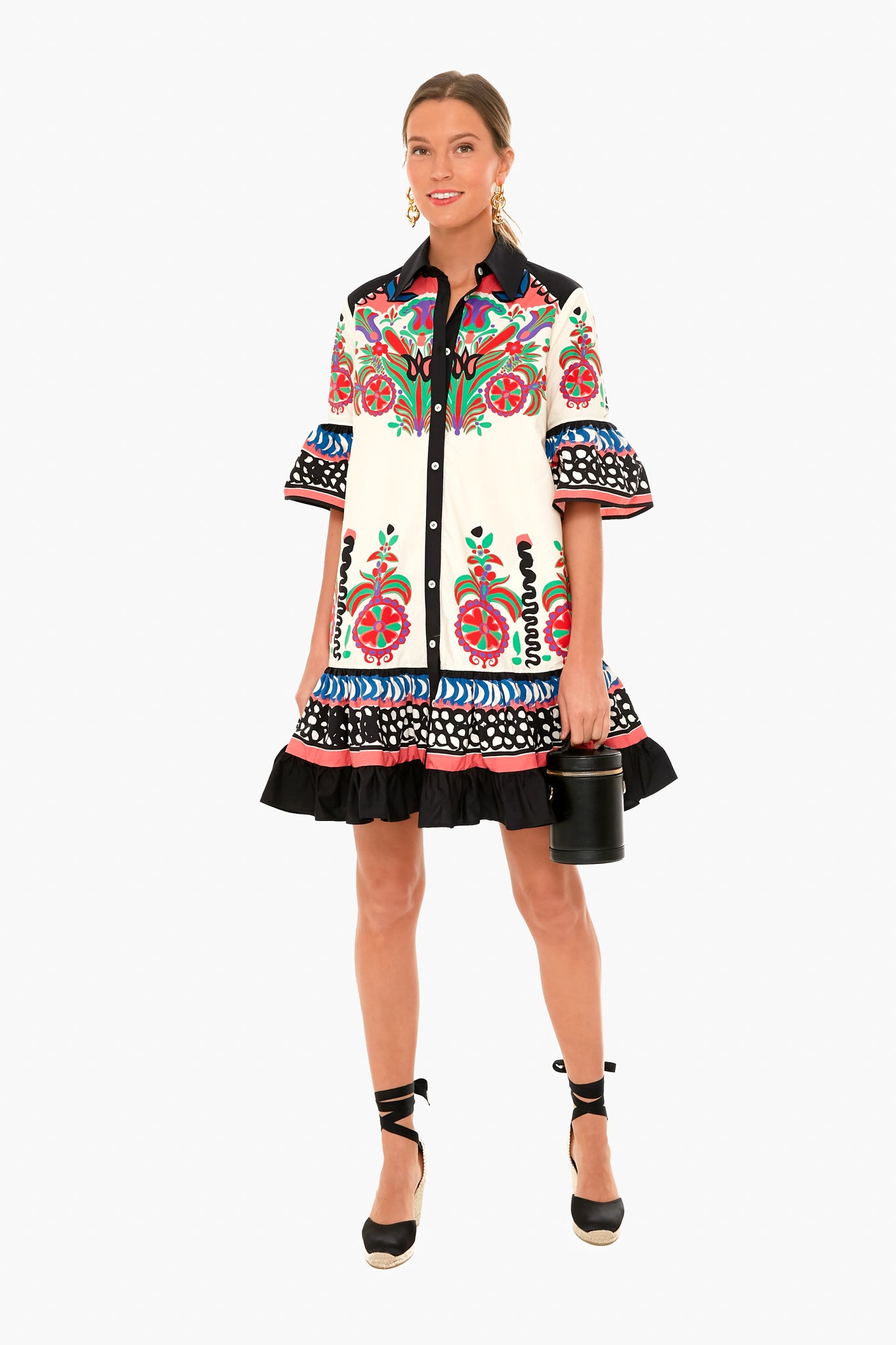 A woman wears a La DoubleJ cotton poplin dress with vibrant floral and geometric prints, black ruffled hem, paired with black espadrille wedges and a small black cylindrical handbag. Made in Italy.