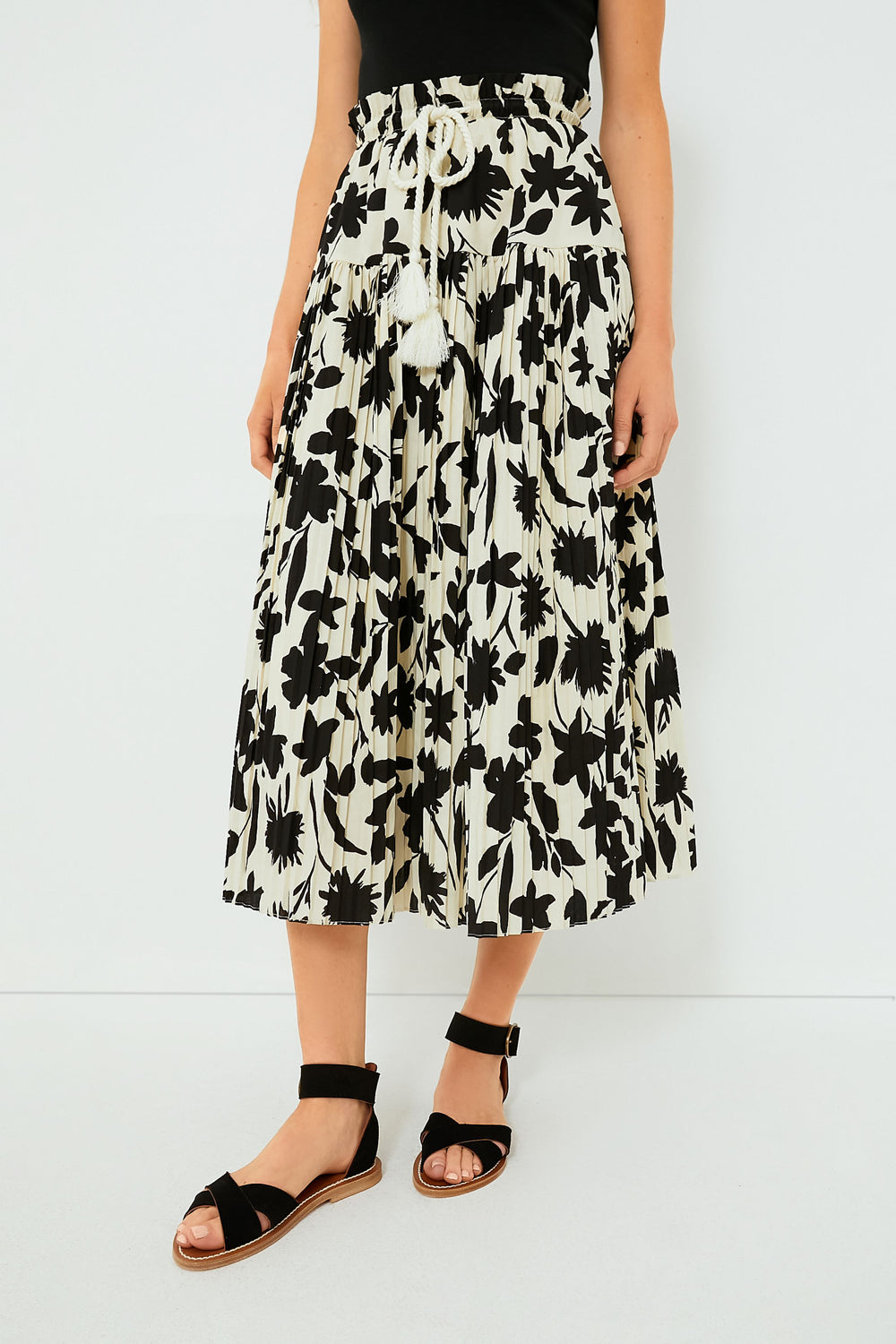 Someone wears a black top, a Moon River Ivory Floral Skirt with a rope belt and ruffled waistband, and black sandals.