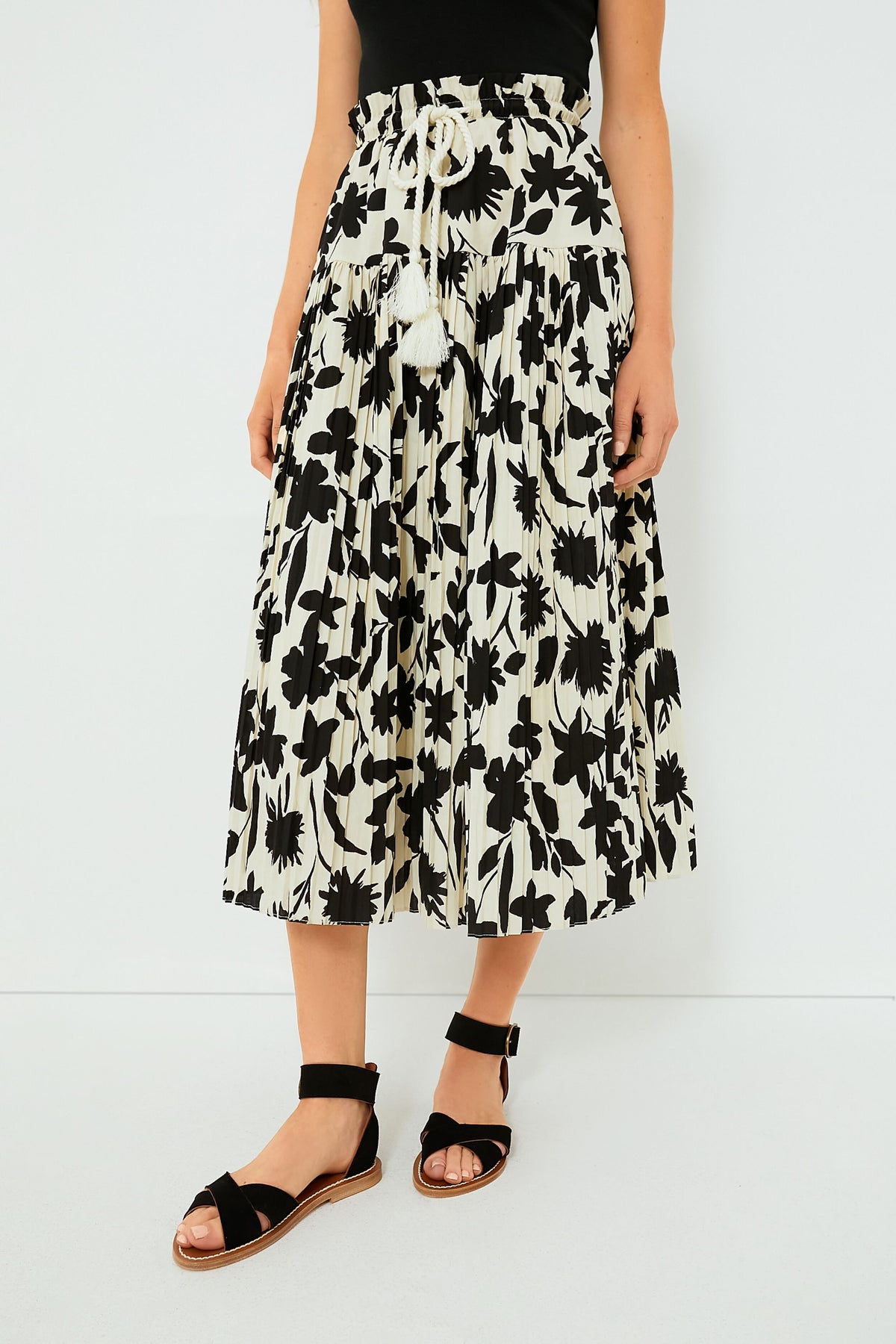 Someone wears a black top, a Moon River Ivory Floral Skirt with a rope belt and ruffled waistband, and black sandals.