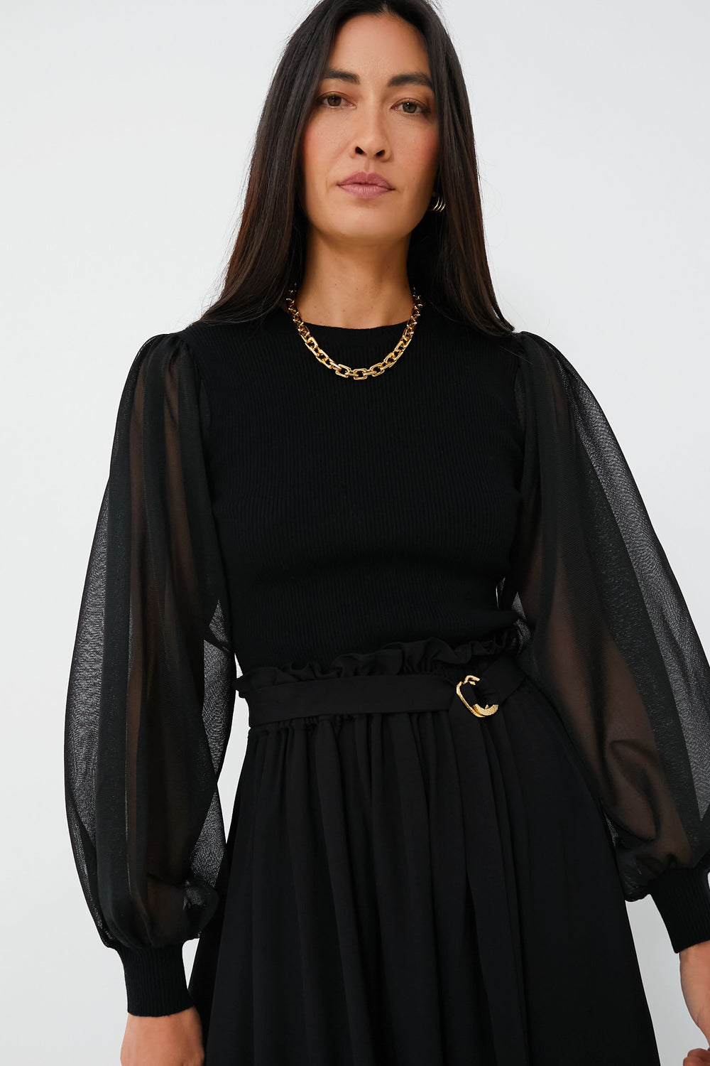 A woman with long dark hair wears an elegant Ulla Johnson black top featuring sheer puff sleeves, a black skirt, a gold chain necklace, and a black belt with a gold buckle.
