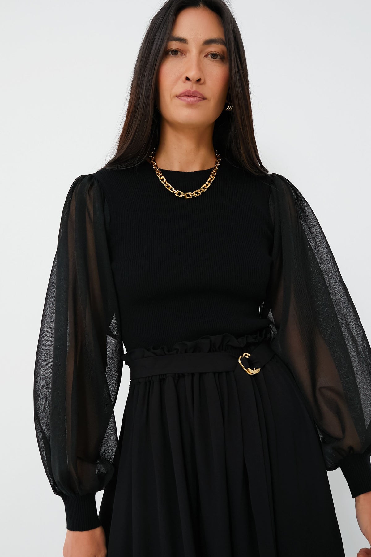 A woman with long dark hair wears an elegant Ulla Johnson black top featuring sheer puff sleeves, a black skirt, a gold chain necklace, and a black belt with a gold buckle.