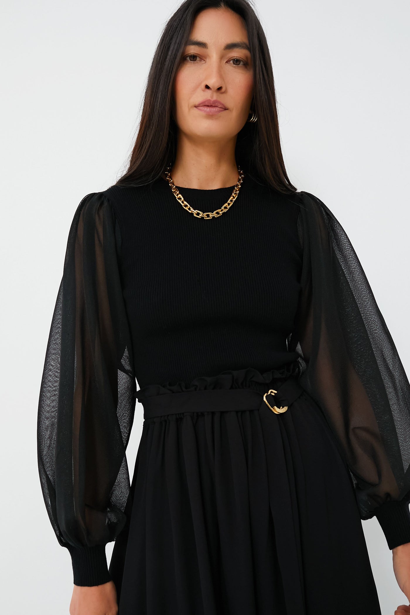 A woman with long dark hair wears an elegant Ulla Johnson black top featuring sheer puff sleeves, a black skirt, a gold chain necklace, and a black belt with a gold buckle.