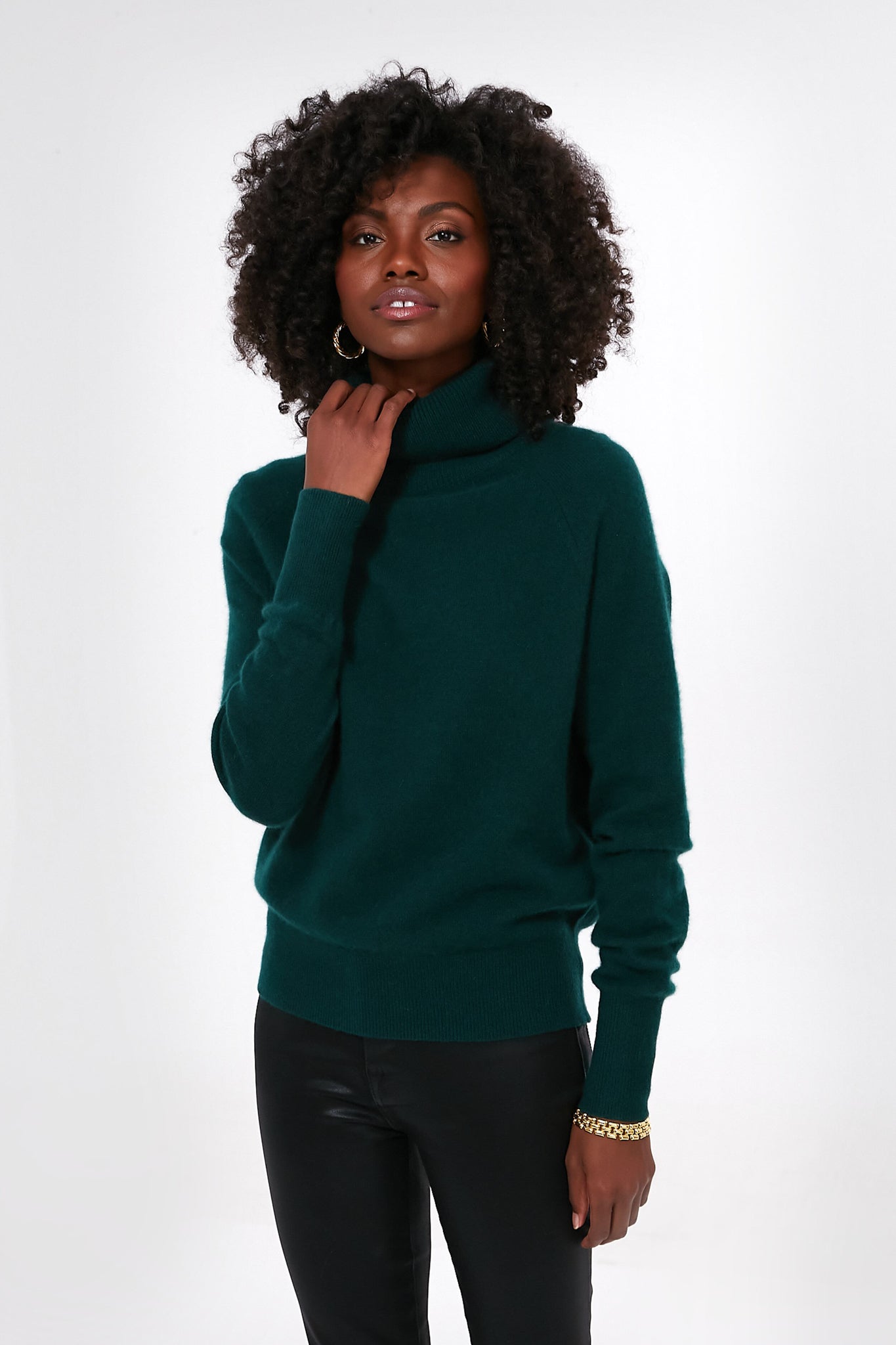 A woman wears a White + Warren Cyprus Green cashmere turtleneck sweater with ribbed trim and black pants.