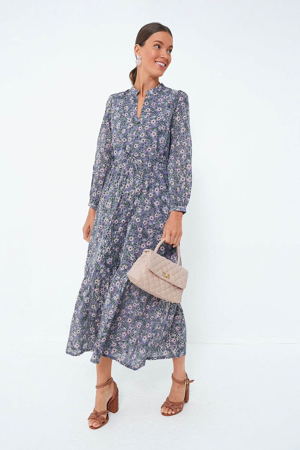 A woman wears a Love Tomas by Emerson Fry long-sleeved floral dress in organic cotton, holding a beige quilted handbag and wearing brown heeled sandals.