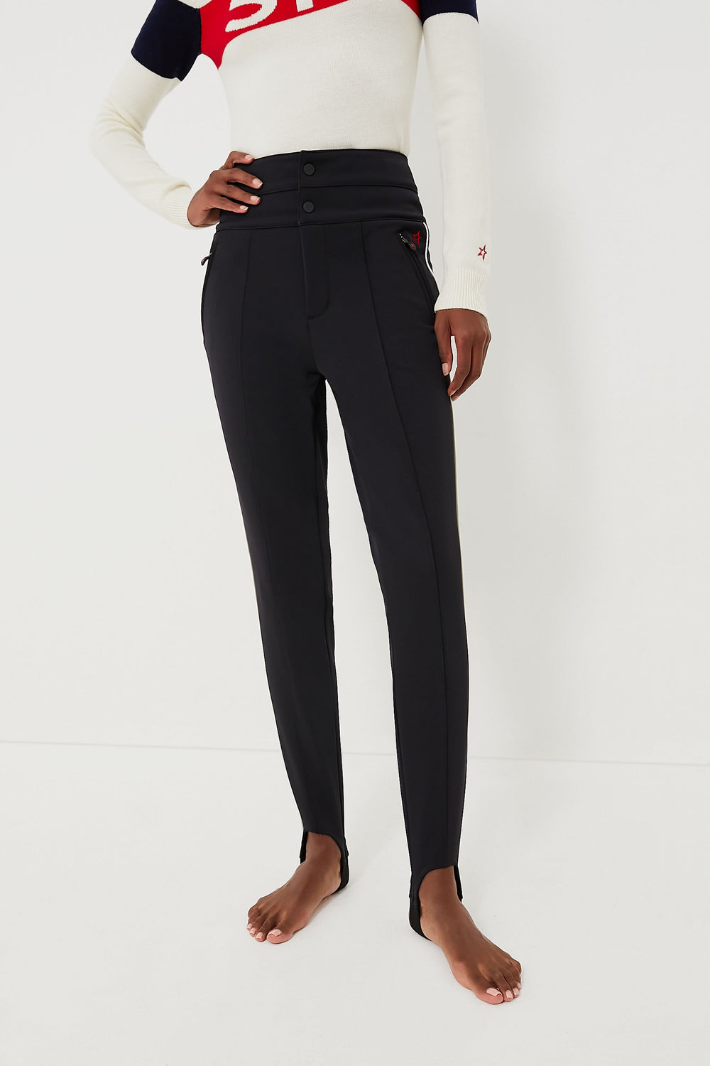 A person stands barefoot in Perfect Moment high-waisted black ski race pants with front buttons and a long-sleeve white, red, and navy top.